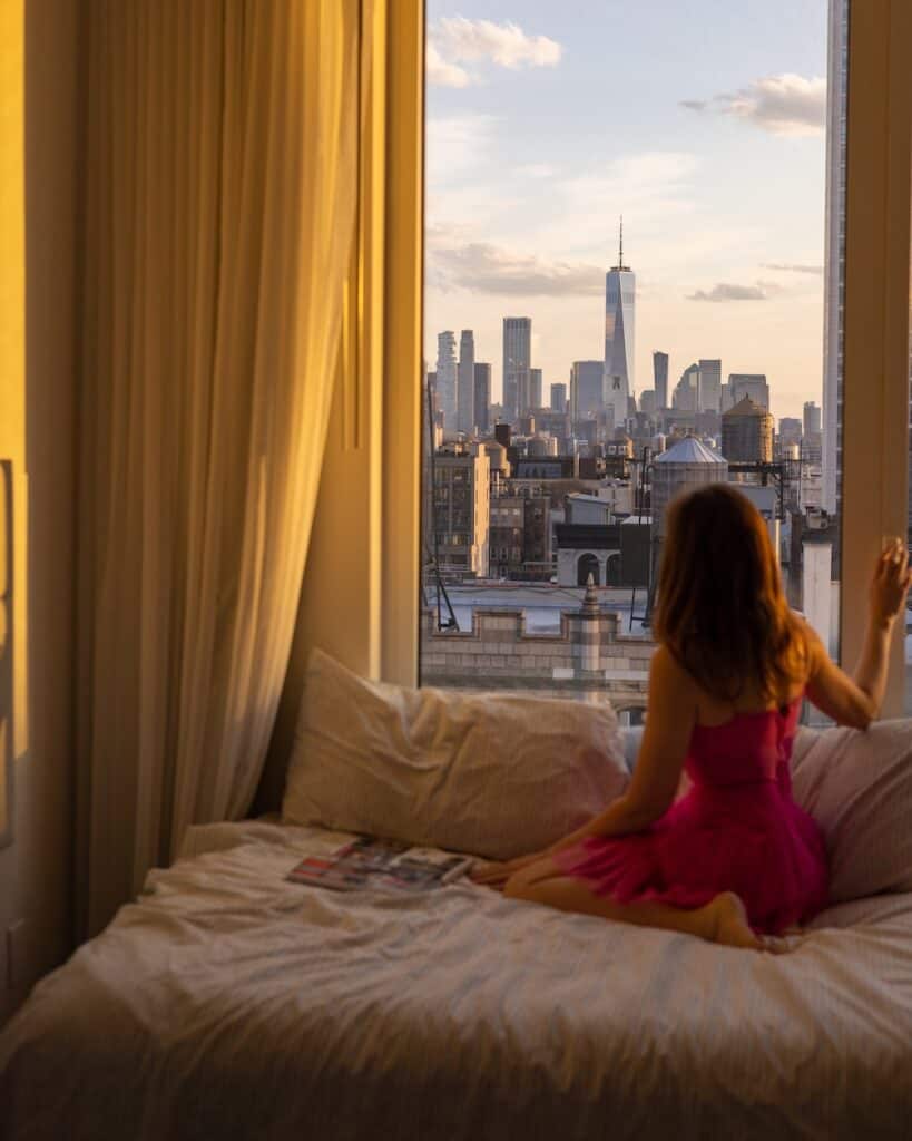 Laura sitting on bed looking at NYC skyline through window, ready to go after her dreams