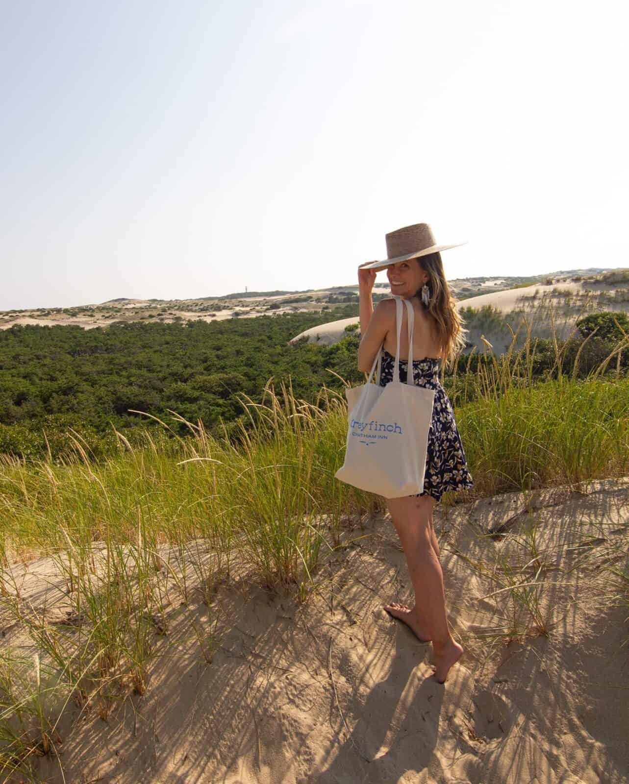 Laura wearing a sun hat and a floral sundress standing barefoot on a sandy crest of the Provincetown sand dunes, carrying a canvas tote bag and looking back over her shoulder at the sweeping green valley below.