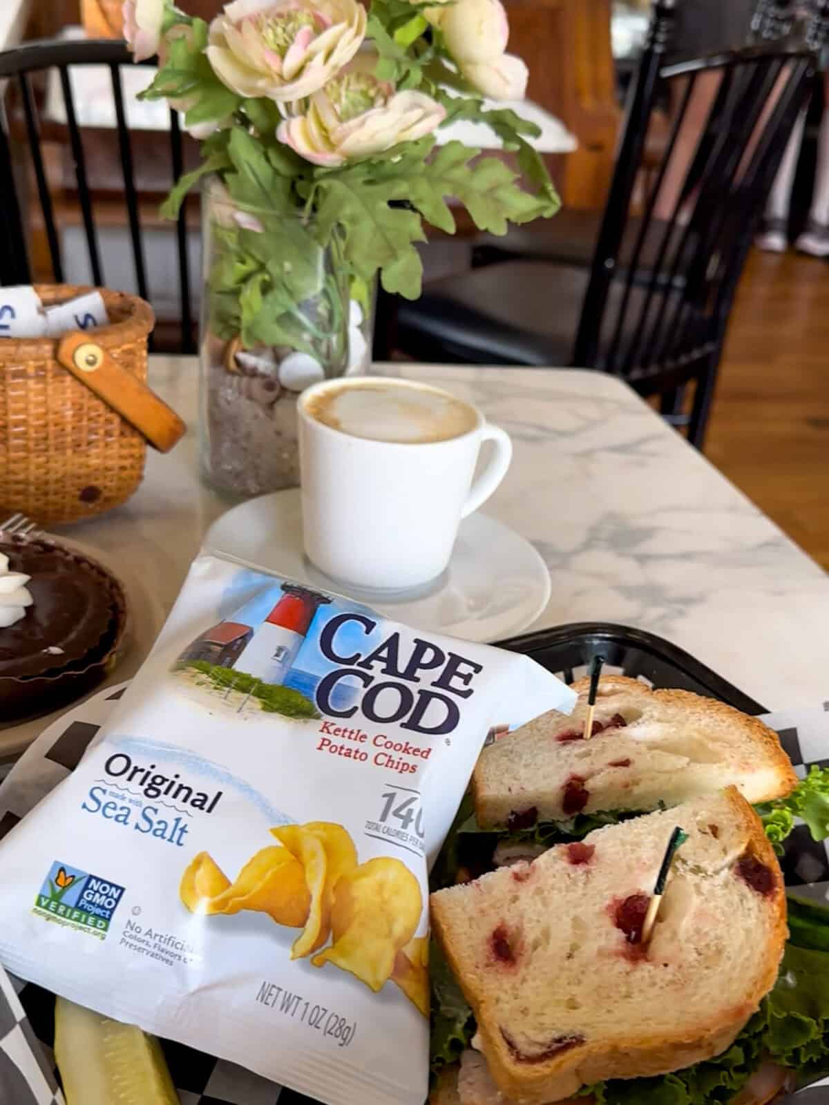 A white marble cafe table holds a fresh turkey and cranberry sandwich on artisanal bread, a bag of Original Cape Cod Kettle Cooked Potato Chips, a white mug of hot cappuccino, and a small vase of flowers.