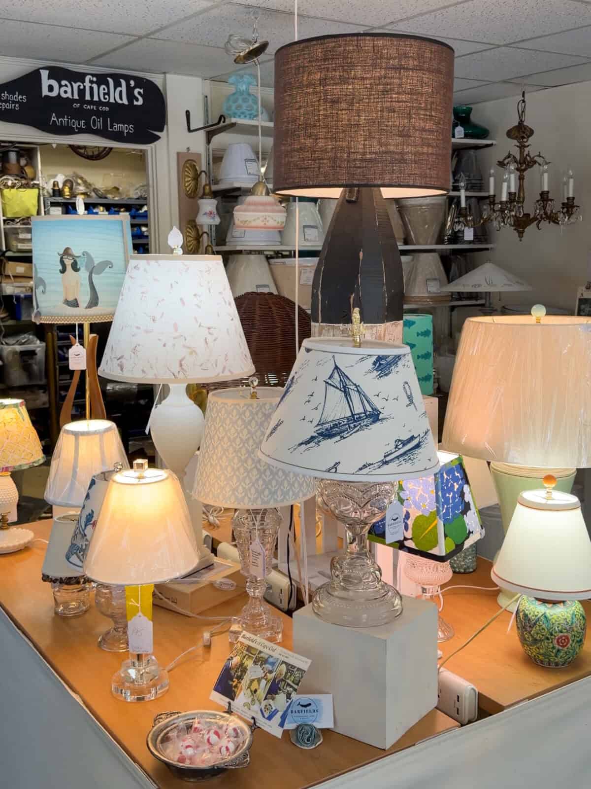 A warm, well-lit tabletop display inside Barfield's showing various glowing lamps, including a central clear glass lamp with a blue-and-white sailboat patterned shade, a tall black rustic lamp, and a whimsical shade decorated with a painted mermaid.