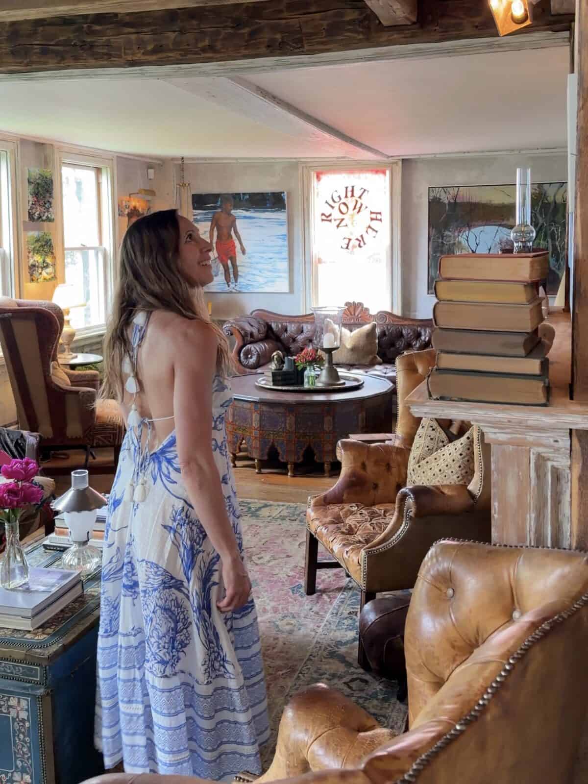 Laura in a blue-and-white patterned halter dress stands smiling in the center of an artistic, sunlit living room with rustic wooden beams, looking up past a stack of antique hardcover books on a mantelpiece toward vibrant paintings decorating the walls.