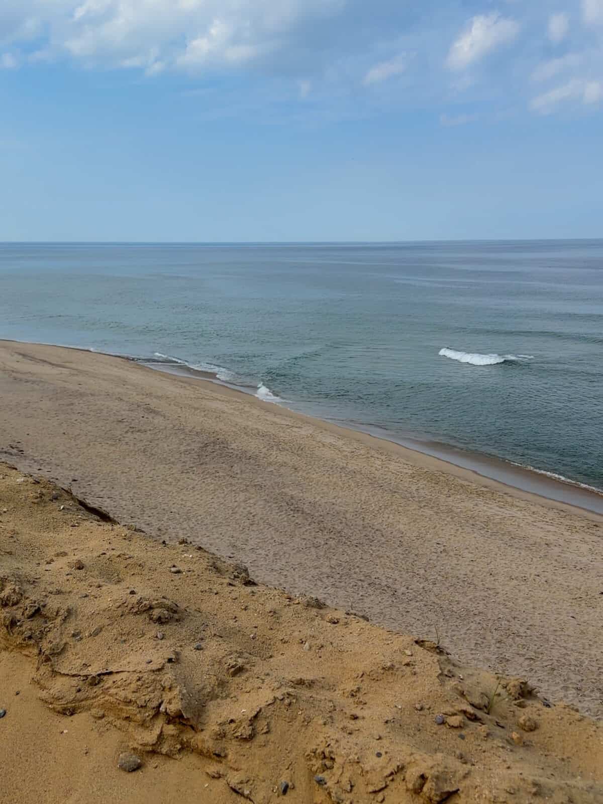 A scenic view looking down from the sandy edge of a high dune overlooking the wide, serene sandy shore of Cahoon Hollow Beach meeting a calm, deep blue Atlantic Ocean under a lightly clouded blue sky.