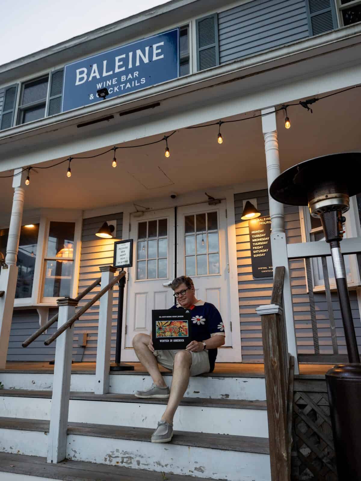 DJ, Braintree Jim sits on the wooden front steps of the grey-shingled Baleine Wine Bar building under glowing string lights, holding a Gil Scott-Heron "Winter in America" vinyl album cover.