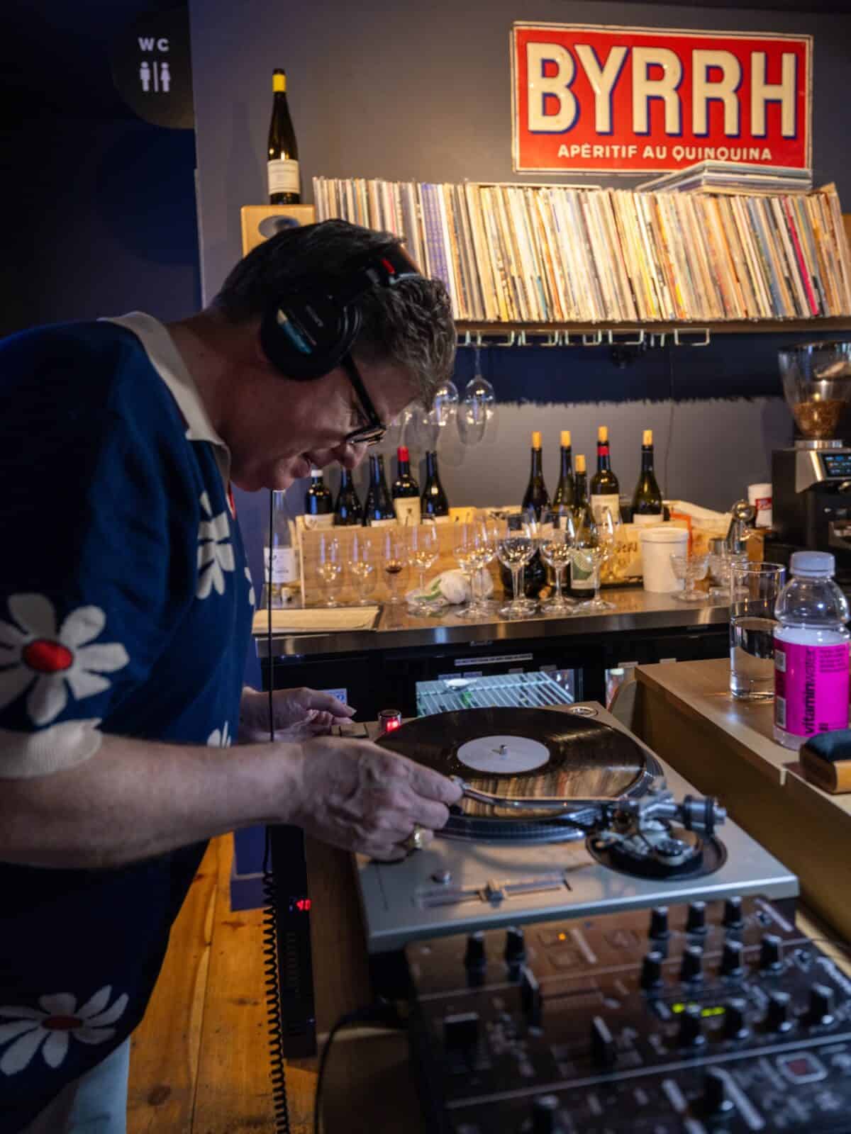 DJ, Braintree Jim wearing headphones and a floral-patterned shirt cues up a vinyl record on a turntable at a cozy bar counter stocked with wine bottles and glasses under a vintage red "BYRRH" aperitif sign.