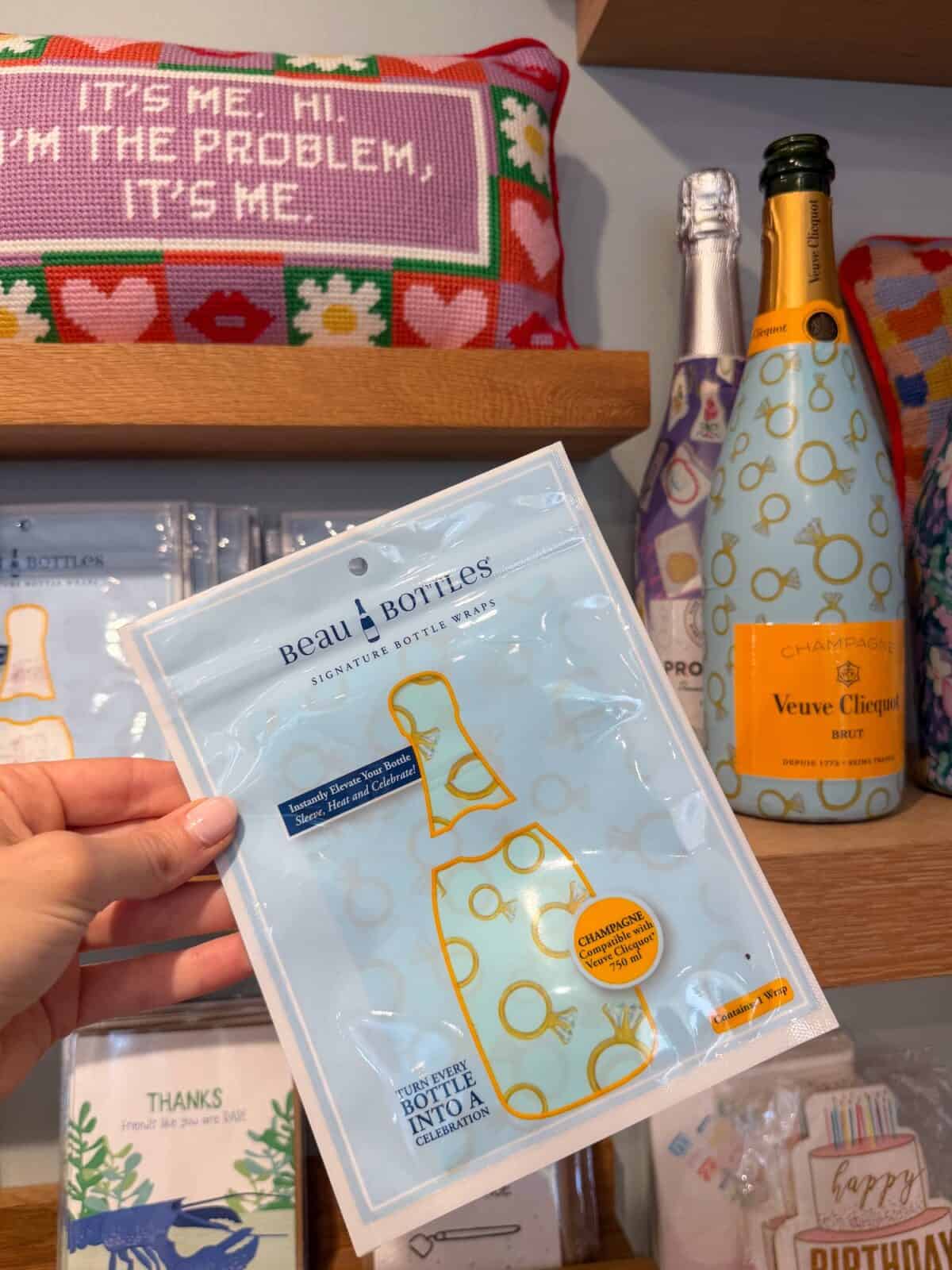 A close-up of a hand holding a blue-and-gold "Beau Bottles Signature Bottle Wrap" designed for champagne bottles, displayed in front of custom-wrapped Veuve Clicquot bottles on a wooden gift shop shelf.