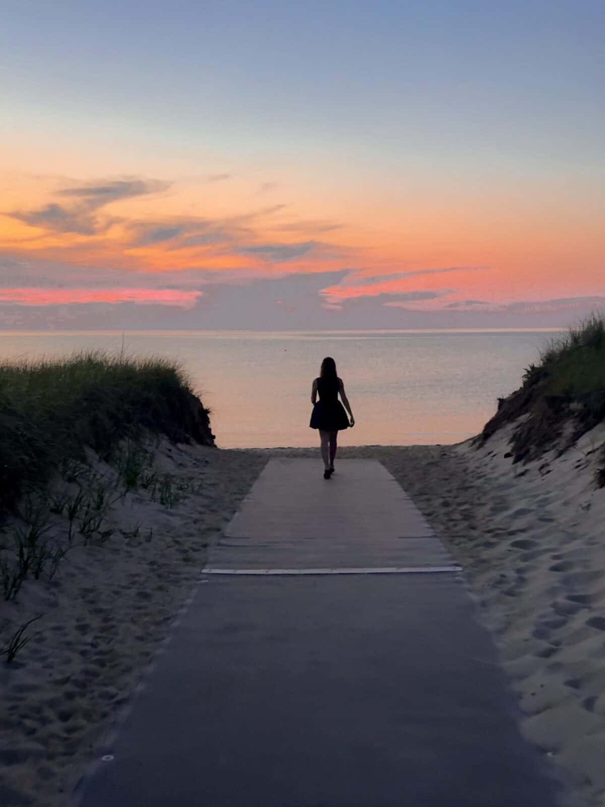 The silhouette of Laura walking down a long beach mat walkway cutting through grassy sand dunes toward a calm ocean during a spectacular, fiery orange and yellow sunset at Breakwater Beach in Brewster.