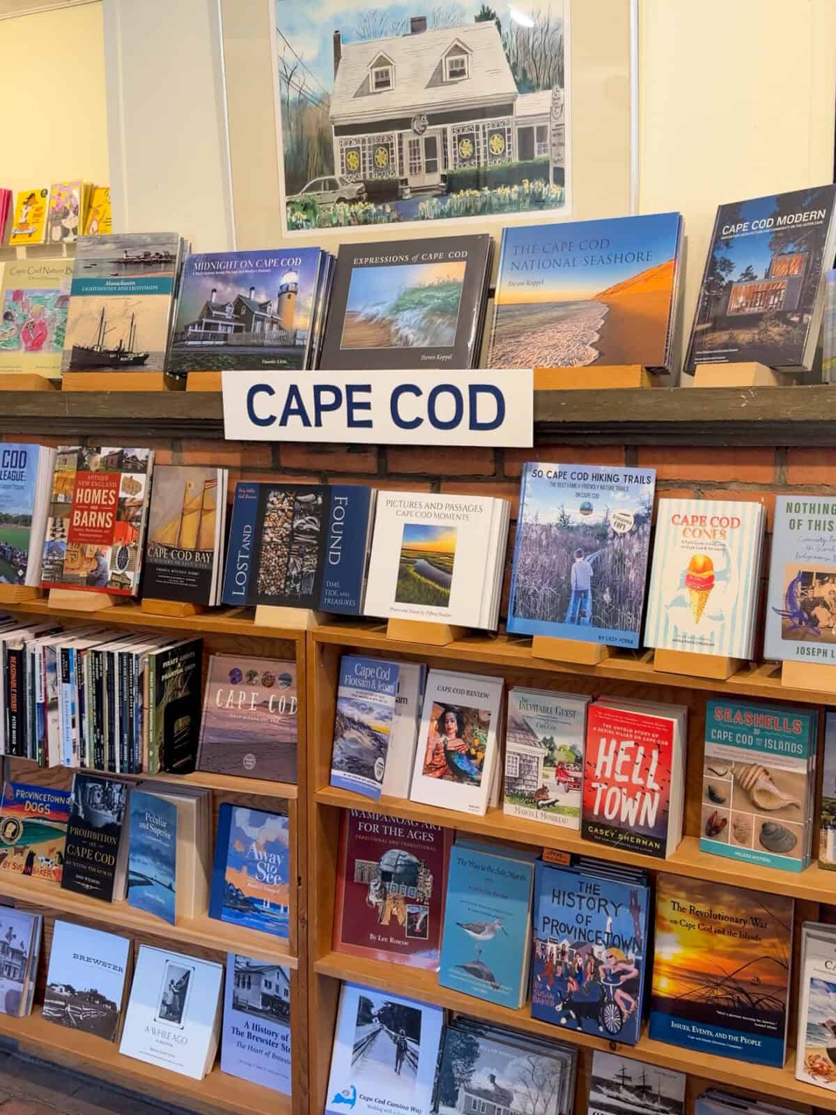 A well-stocked wooden bookshelf dedicated to Cape Cod local interest books, featuring titles like The Cape Cod National Seashore, 50 Cape Cod Hiking Trails, Hell Town, and The History of Provincetown beneath a painted sign labeled "CAPE COD".