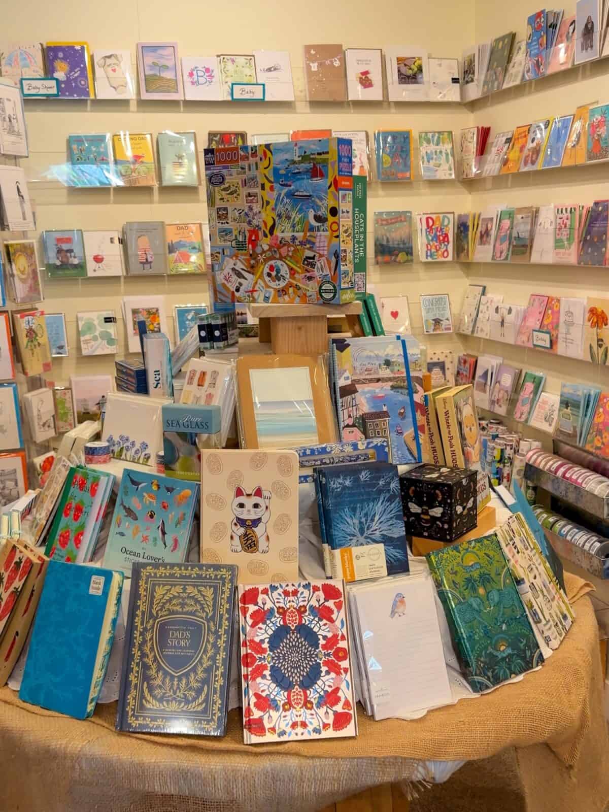 A colorful circular display table inside a bookstore covered in burlap cloth, showcasing patterned journals, a lucky cat notebook, stationery kits, wrapped greeting cards lining the walls, and a large seaside puzzle box at the center.