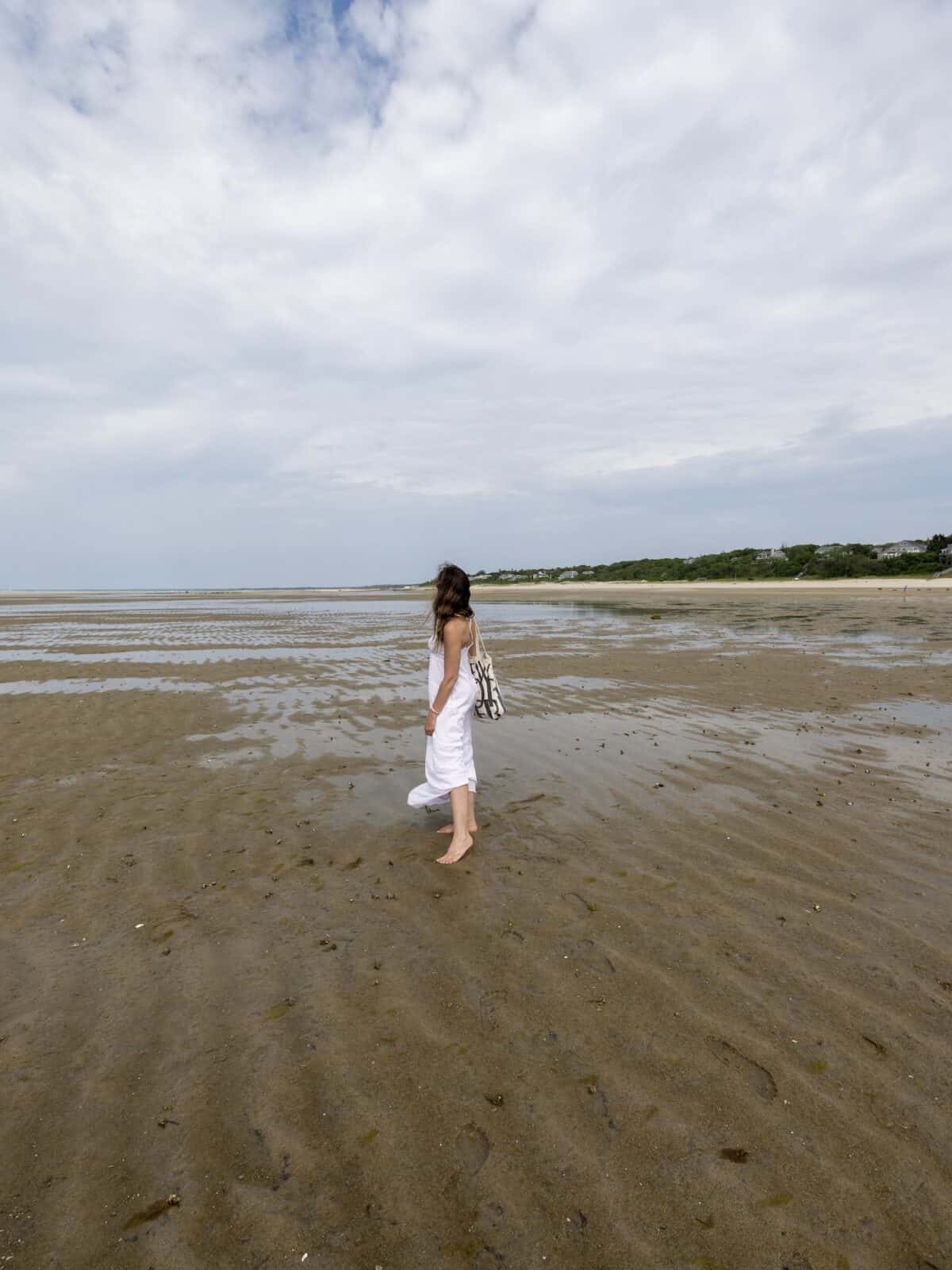 Laura in a long white summer dress stands barefoot on the wet, rippled sandbars of the Brewster Flats during low tide under a dramatic cloudy sky, looking out across the vast expanse towards the shoreline.