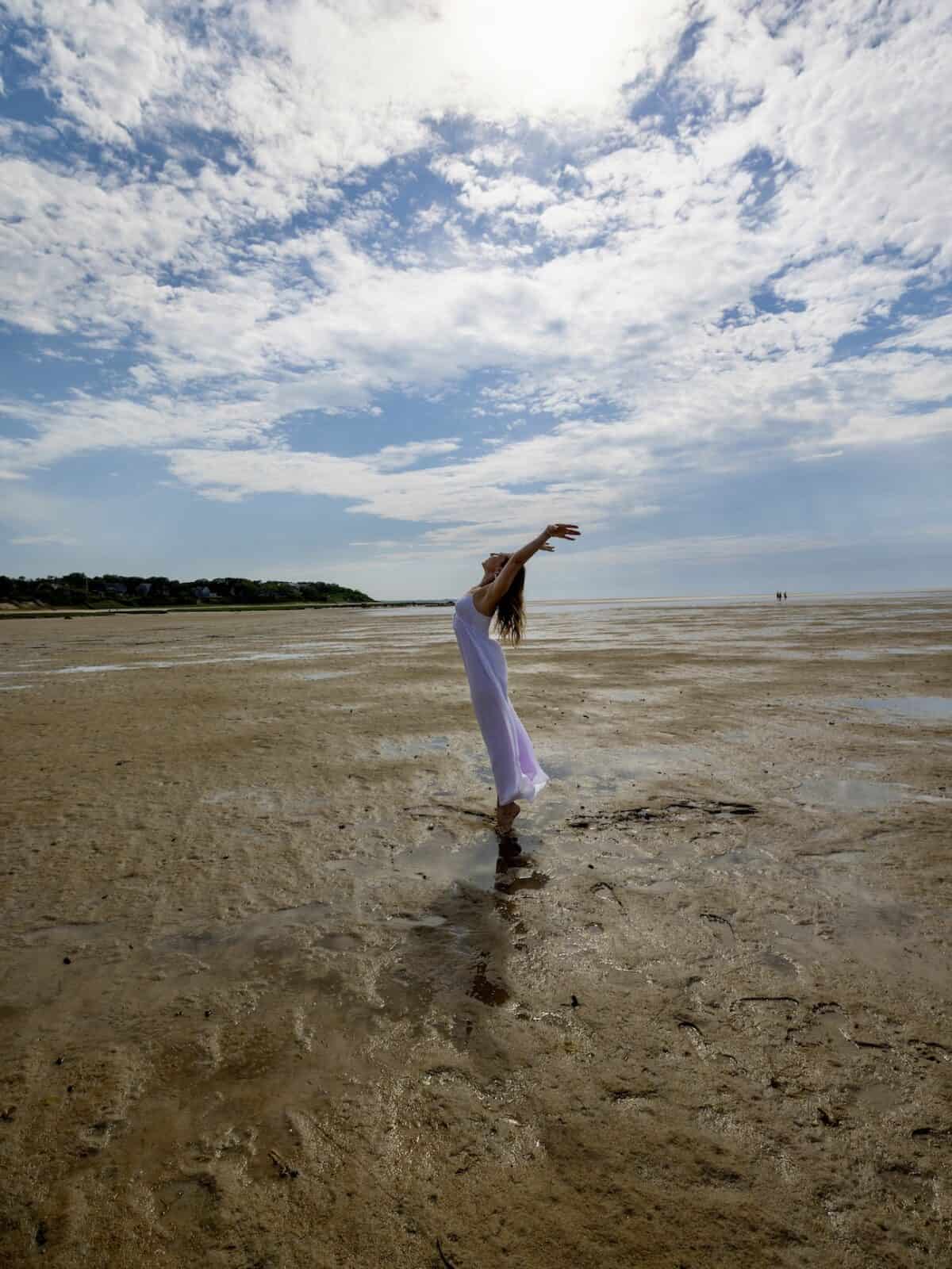 Laura wearing a white dress stands on the reflective wet sand of the tidal flats, leaning back gracefully with her arms stretched up toward a sun-drenched sky filled with wispy blue and white clouds.