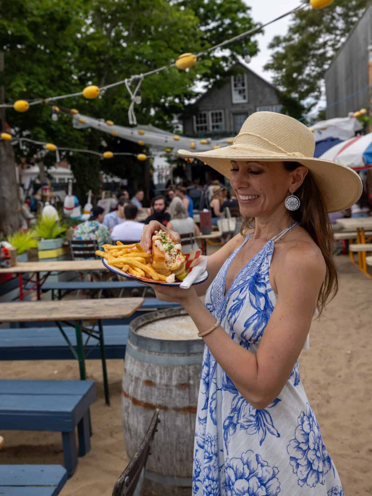 Laura in a wide-brimmed straw hat and a blue-and-white floral dress stands in a sandy backyard patio courtyard, admiring a plate with a fresh lobster roll and fries at The Canteen in Provincetown, underneath a string of yellow festive lights.