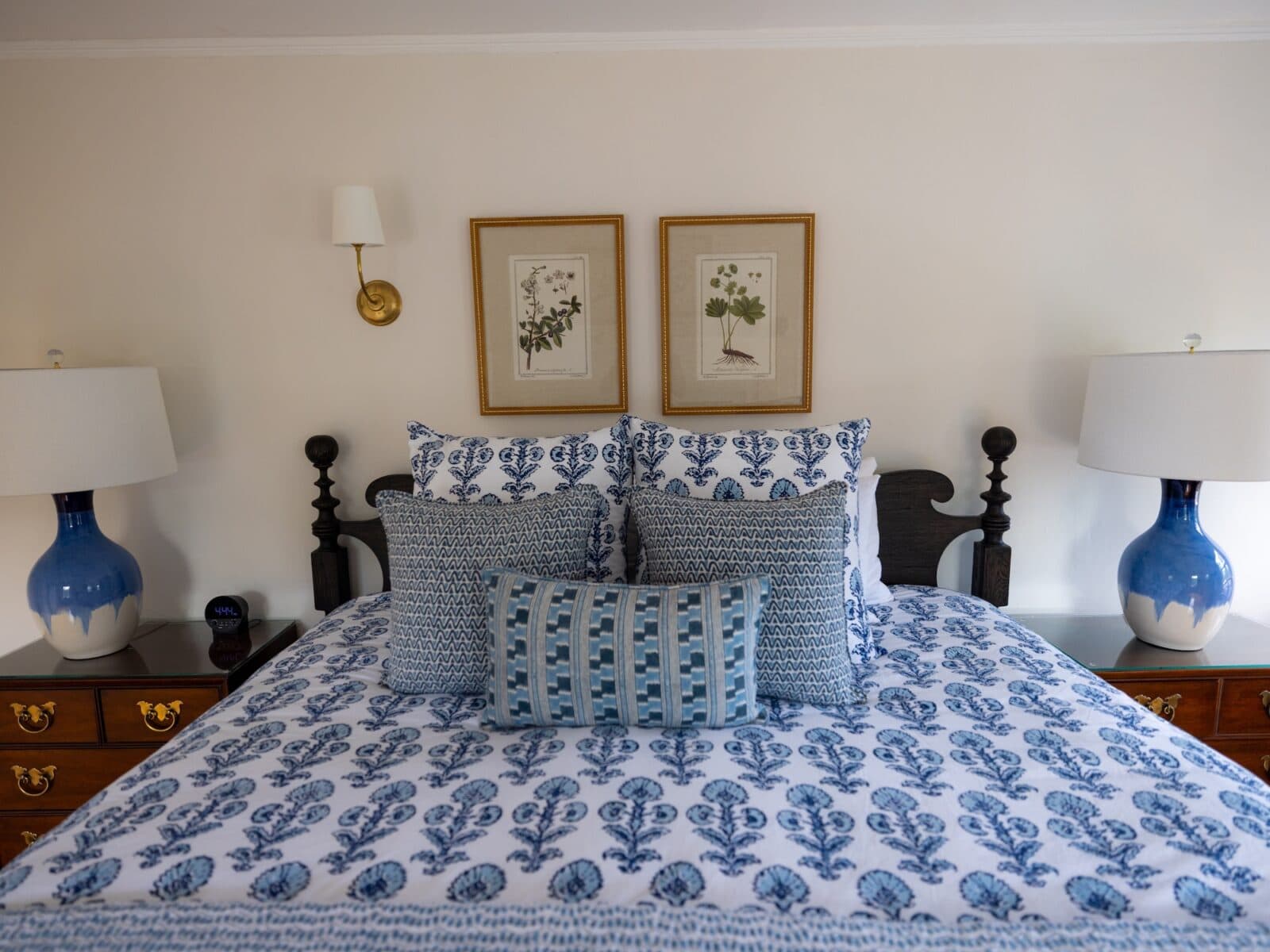 Symmetrical view of a luxury bed at Charm on Main with crisp blue and white block-print pattern bedding, matching decorative throw pillows, two blue ceramic bedside lamps on wooden nightstands, and twin botanical art prints framed on the wall.