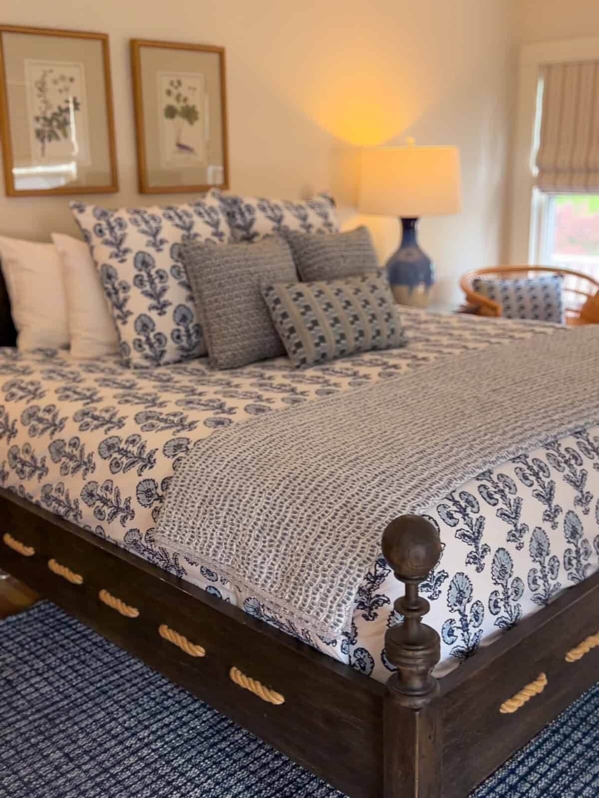 A cozy corner view of a deluxe bedroom at Charm on Main, showcasing a dark wooden bed frame accented with nautical rope details, styled with blue and white pattern linens, a lit bedside lamp, and a blue woven area rug.
