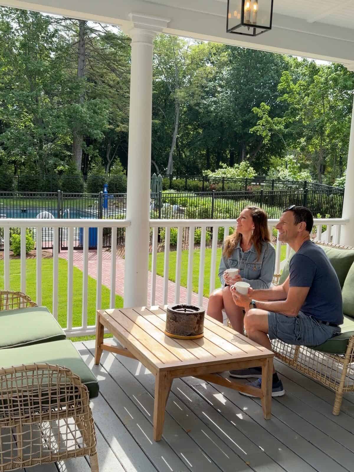 Laura and Luca sit with coffee cups on a covered white porch at Charm on Main, relaxing on outdoor furniture with green cushions while looking out at the swimming pool fence and mature green trees in the backyard.
