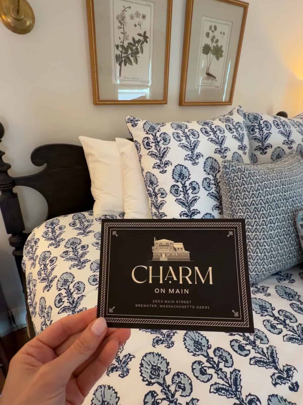 Laura holds up a black and white welcome card featuring the "Charm on Main" logo and their address in Brewster, Massachusetts, held in front of a luxury bed styled with blue patterned pillows and a dark wood headboard.