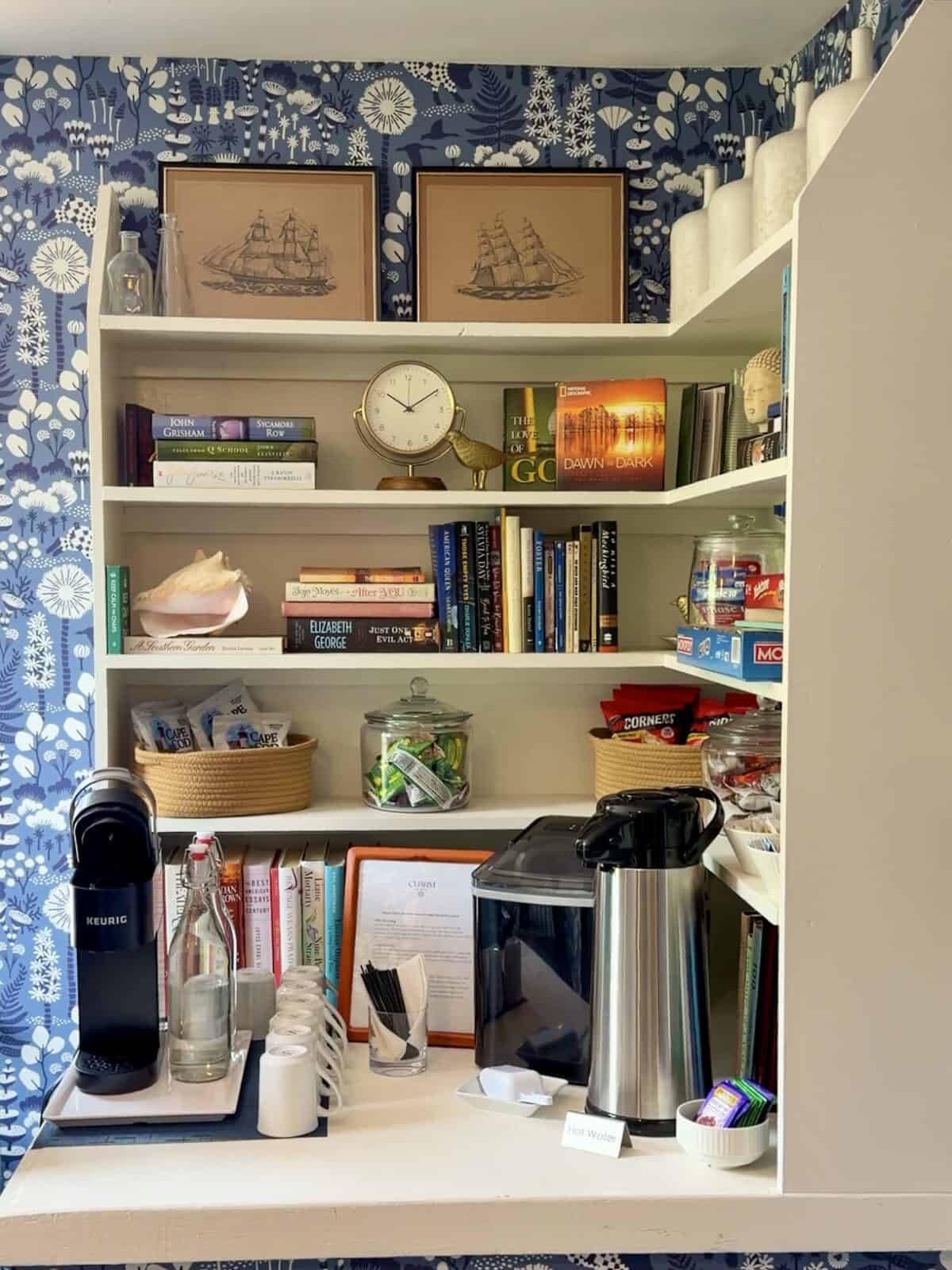A beautifully styled multi-tiered white corner built-in bookcase serving as a guest hospitality coffee and snack nook inside the Charm bed and breakfast in Provincetown, decorated with bold blue-and-white botanical pattern wallpaper, nautical sailing ship artwork, framed hotel info, a Keurig coffee maker, snack baskets with Cape Cod potato chips, and neatly stacked books