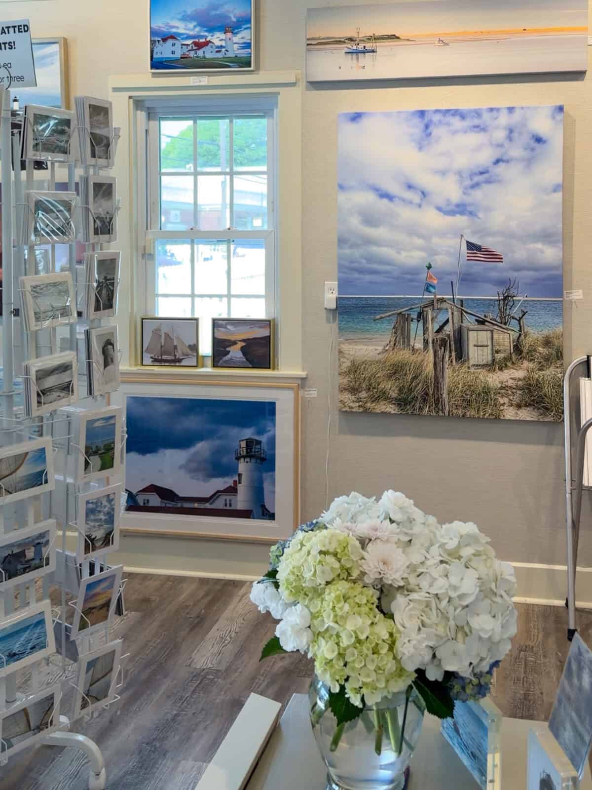 Interior of a coastal art gallery in Chatham featuring framed prints of New England lighthouses, coastlines, and sailboats, with a lush vase of white and green hydrangeas sitting on a table in the foreground.
