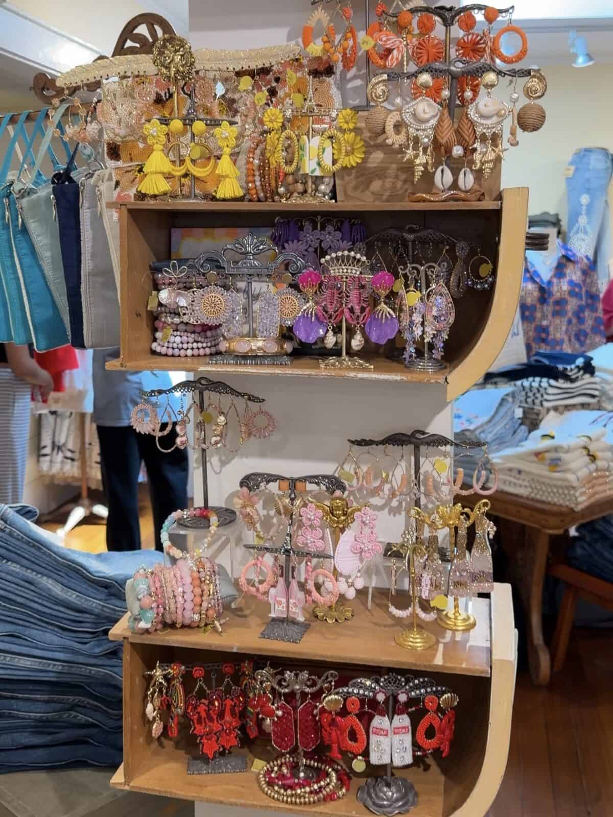 A multi-tiered wooden shelf displaying rows of intricate, handmade bohemian-style earrings in vibrant shades of pink, yellow, purple, and coral, alongside stacks of beaded bracelets and folded denim clothing.
