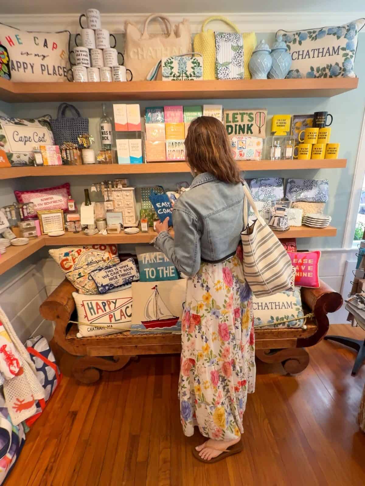 in a denim jacket and floral maxi dress stands inside a coastal boutique, browsing shelves filled with Chatham-themed throw pillows, pastel tote bags, custom coffee mugs, and decorative home accessories.