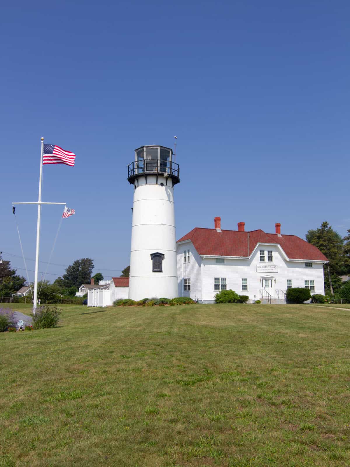A crisp, full-view daytime shot of the white brick Chatham Lighthouse standing tall on a grassy hill next to the white-shingled US Coast Guard Station building with its red roof. An American flag flies prominently on a flagpole under a clear blue sky.