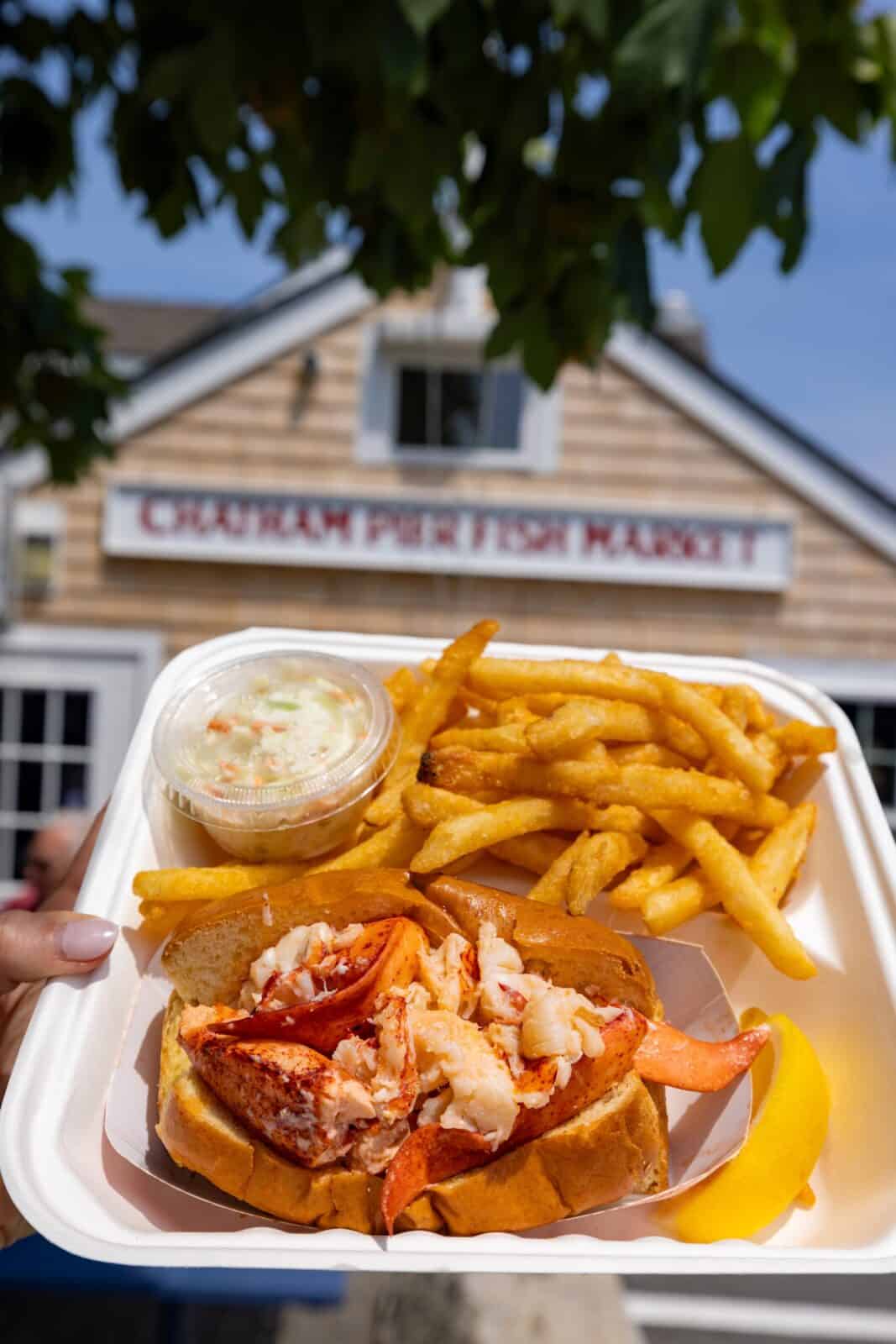 A hand holds up a white container filled with a premium New England lobster roll bursting with fresh lobster meat, served with a side of crispy golden french fries, coleslaw, and a lemon wedge, framed in front of the rustic shingle building of Chatham Pier Fish Market.