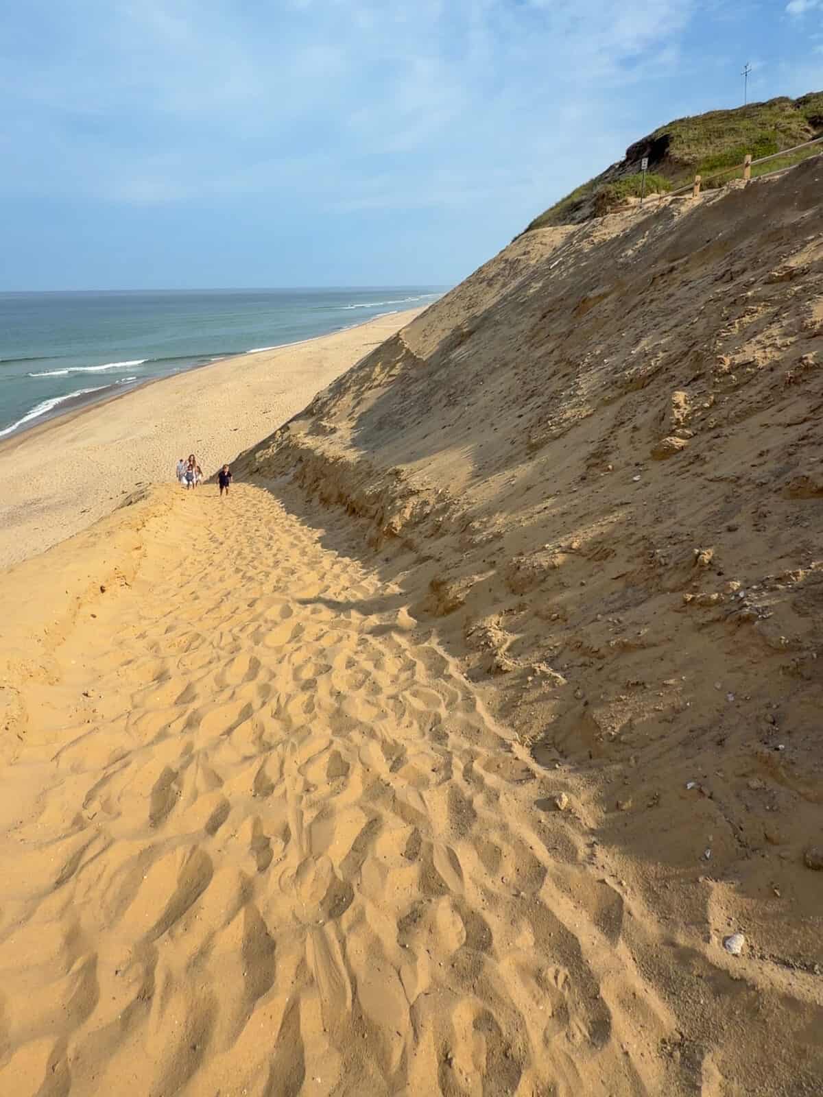 A steep, footprint-laden sand trail climbing up the towering coastal bluff away from the shoreline at Cahoon Hollow Beach in Wellfleet, showing a few small figures near the bottom and a wooden fence along the crest.