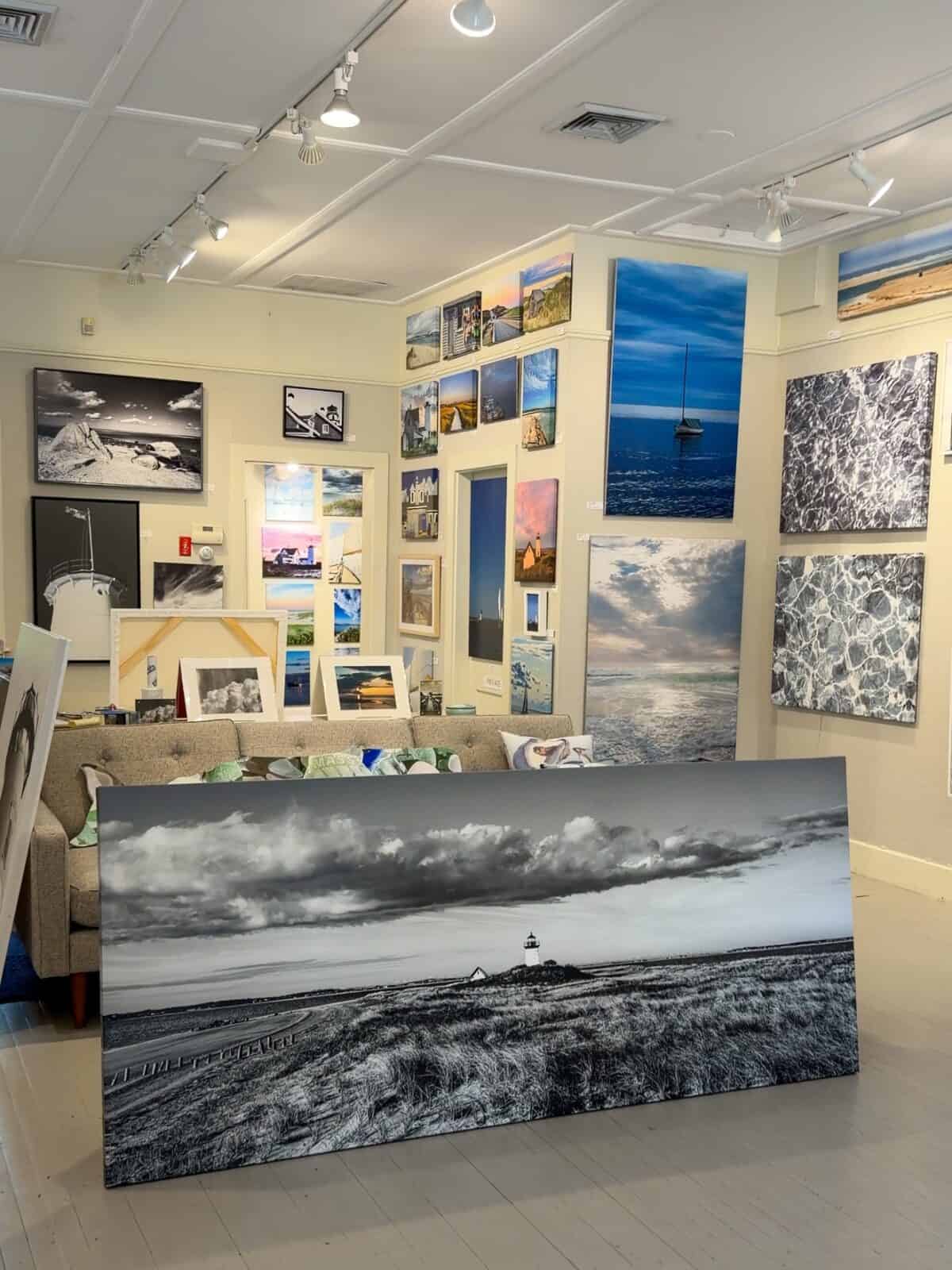 A spacious art gallery showroom in Chatham filled with large canvas prints depicting ocean horizons, lighthouses, and abstract water ripples, centered around a large black-and-white panoramic photograph of a grassy coastal dune resting on the floor.
