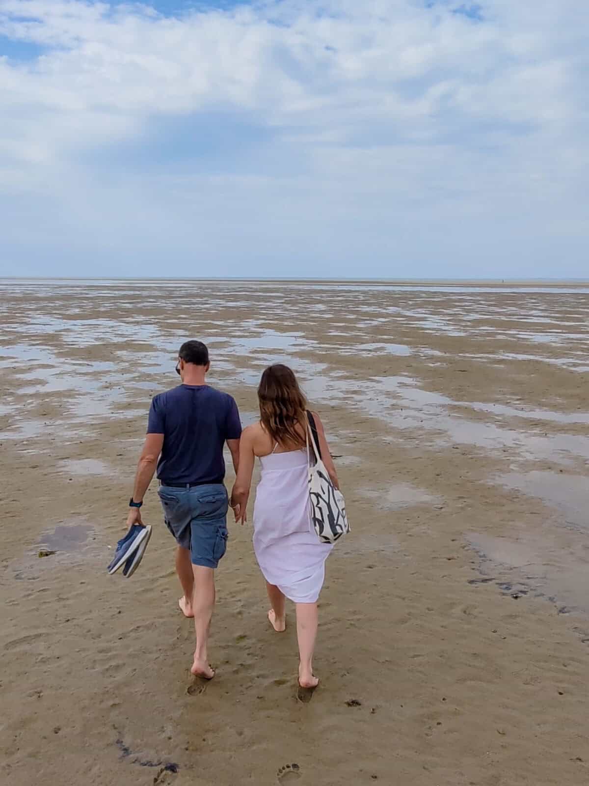 Laura and Luca viewed from behind walks barefoot hand-in-hand out onto the shallow tidal pools of the Brewster Flats; the man carries his shoes in his hand while the woman wears a white dress and carries a patterned tote bag.