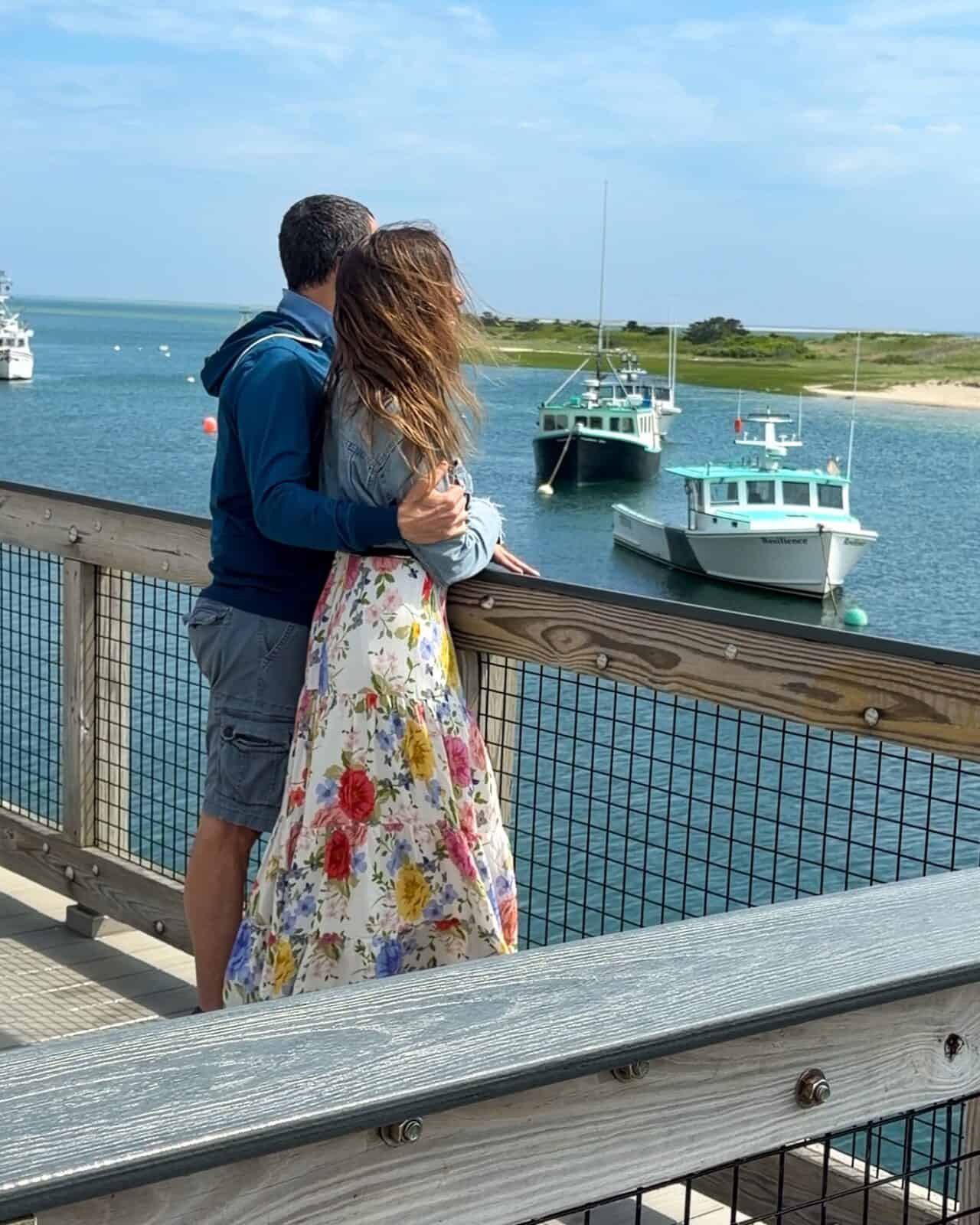 A rearview perspective of Luca in a blue hoodie hugging Laura in a floral maxi dress from behind as they stand together on the wooden railing of Chatham Pier, looking out over commercial fishing vessels docked in the harbor under a sunny sky.