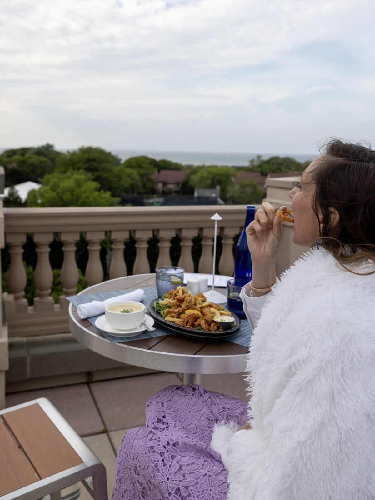 side profile view of Laura wearing a white shaggy jacket and a purple lace dress tasting a piece of crispy fried calamari from her table at Ocean Terrace, with the green trees and blue horizon of Cape Cod Bay blurred in the background.