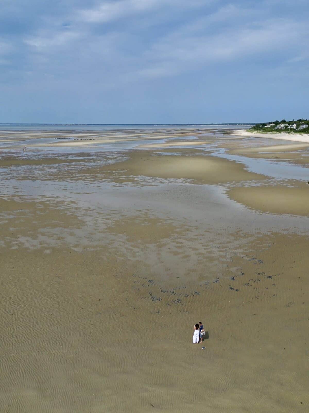 A high-altitude drone shot looking down at a tiny couple standing on an immense expanse of rippled sandbanks during low tide, with coastal luxury homes visible along the distant green bluffs.