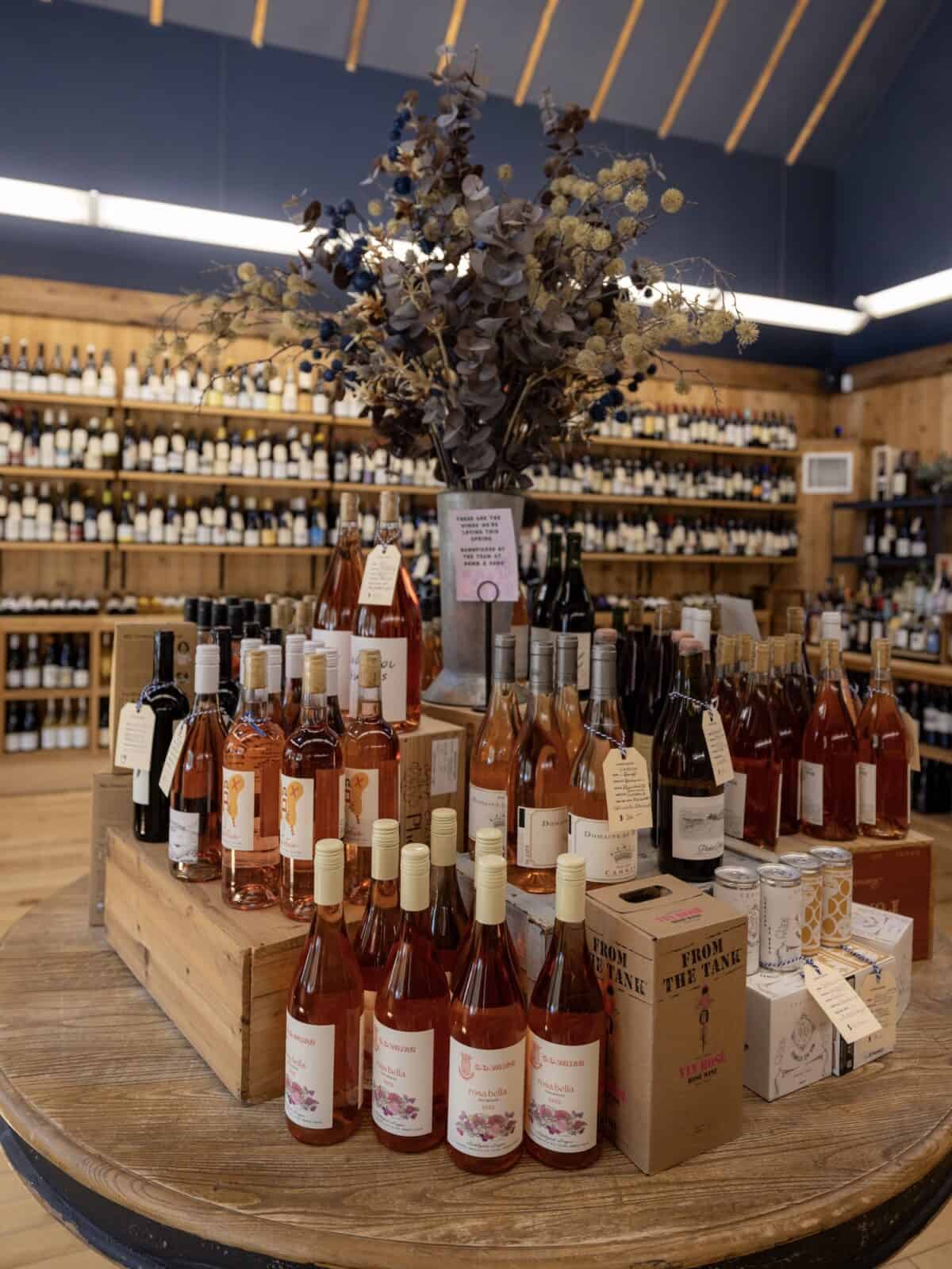 A large, round wooden table in a wine shop features a prominent spring display of rosé and natural wines arranged on wooden crates, centered beneath a large arrangement of dried flowers and eucalyptus.