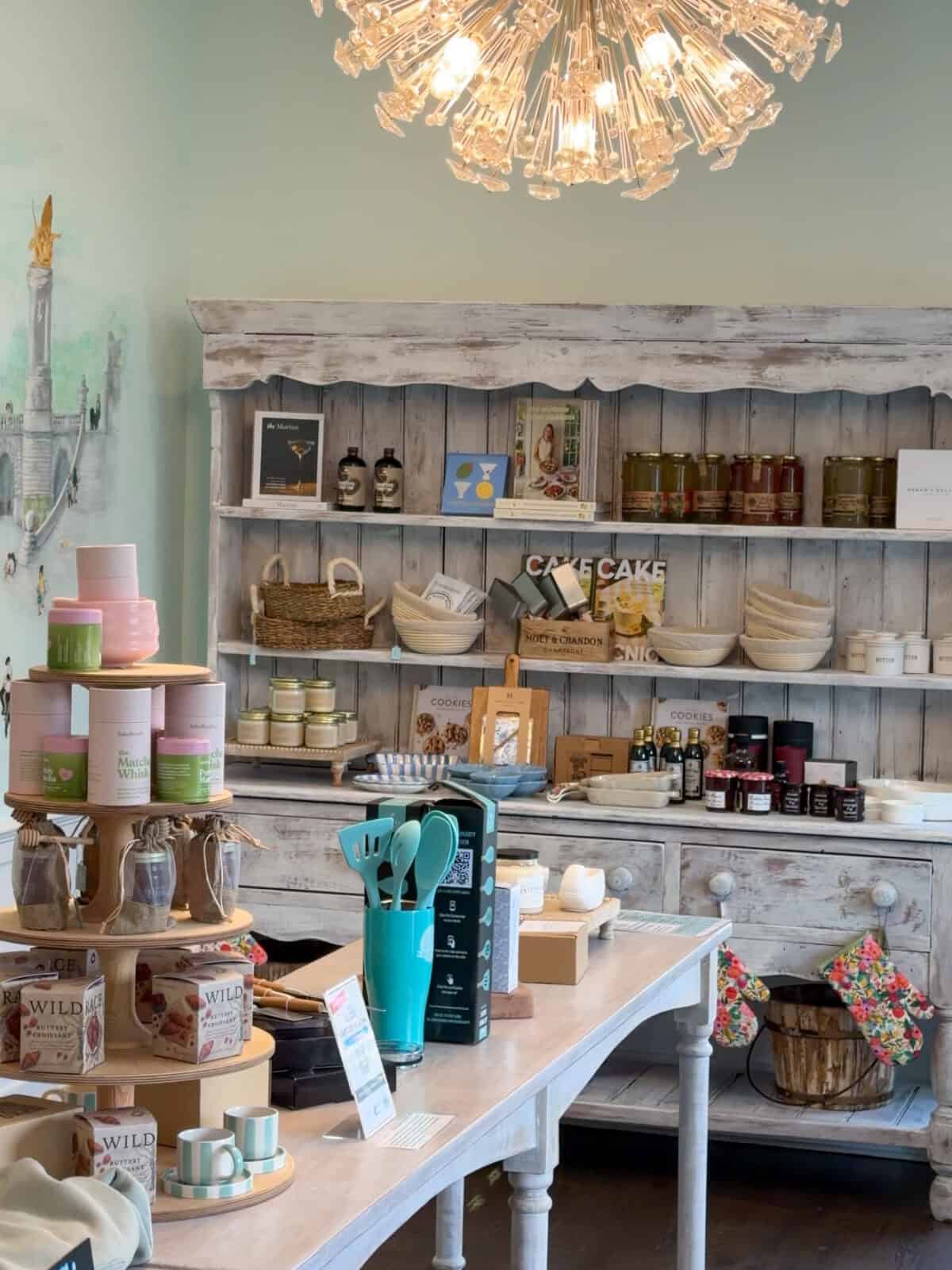 A beautifully styled retail display area inside Eat Cake 4 Breakfast bakery in Brewster, featuring a large rustic white wooden hutch filled with gourmet ingredients, cookbooks, and baking supplies beneath a sparkling modern crystal chandelier, with a display table showcasing colorful tea whisks and pastry boxes in the foreground.