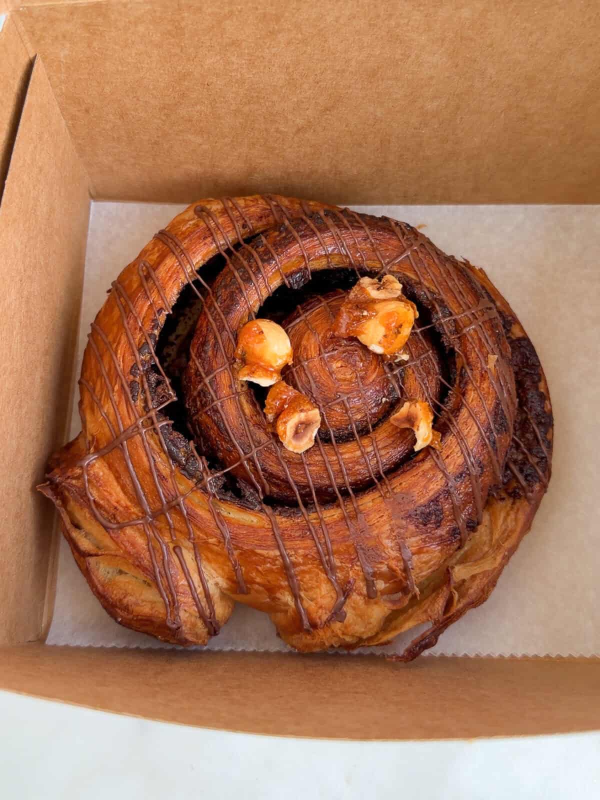 A detailed, top-down close-up shot of a large, perfectly golden-brown laminated spiral pastry drizzled with Nutella and topped with roasted hazelnuts, nestled neatly inside a brown cardboard bakery takeout box.