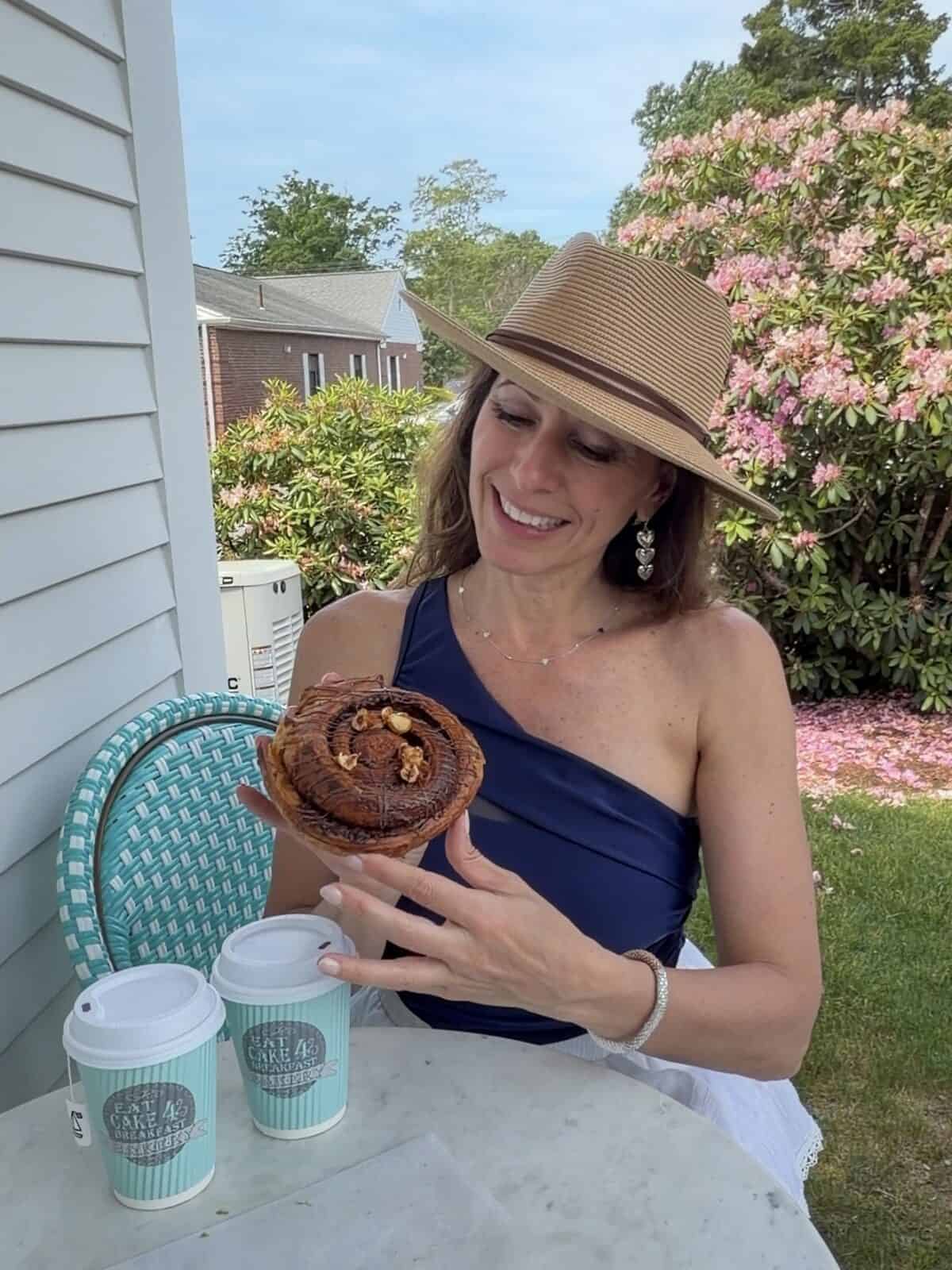 Laura wearing a straw sun hat, a navy blue one-shoulder top, and a white skirt sits at an outdoor marble table on a garden patio, happily holding up a large chocolate-drizzled spiral pastry next to two turquoise "Eat Cake 4 Breakfast" paper coffee cups.