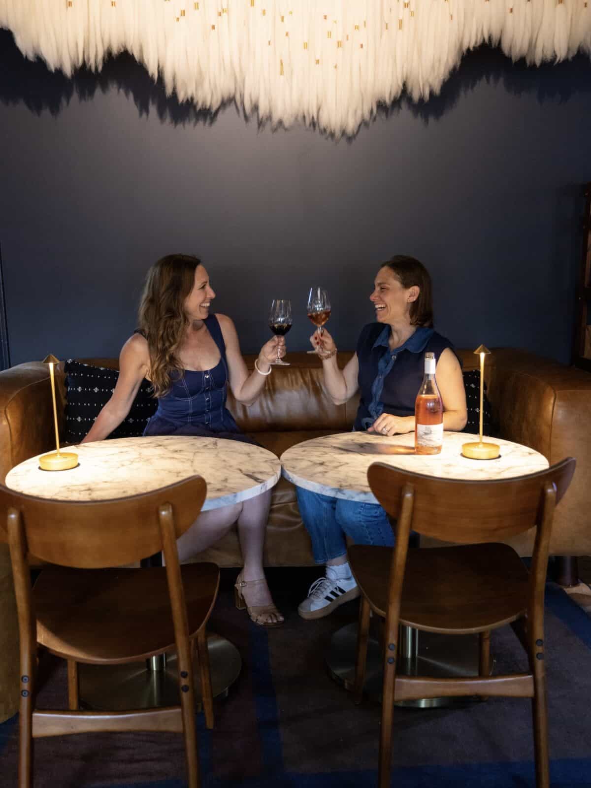 Laura (travel writer) and Erica (owner) sit smiling on a brown leather sofa, raising a glass of red wine and a glass of rosé to toast each other at marble-topped tables inside a stylish dimly-lit lounge with a blue wall.