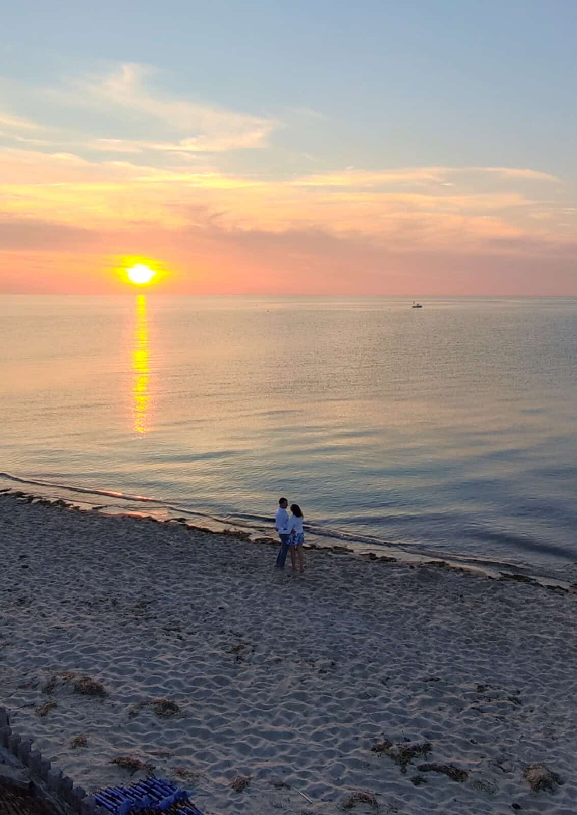 A wide-angle view of a couple standing hand-in-hand on a wide, sandy beach at low tide during a serene pastel yellow and orange sunset, with the sun's reflection creating a long golden path across the smooth ocean water.