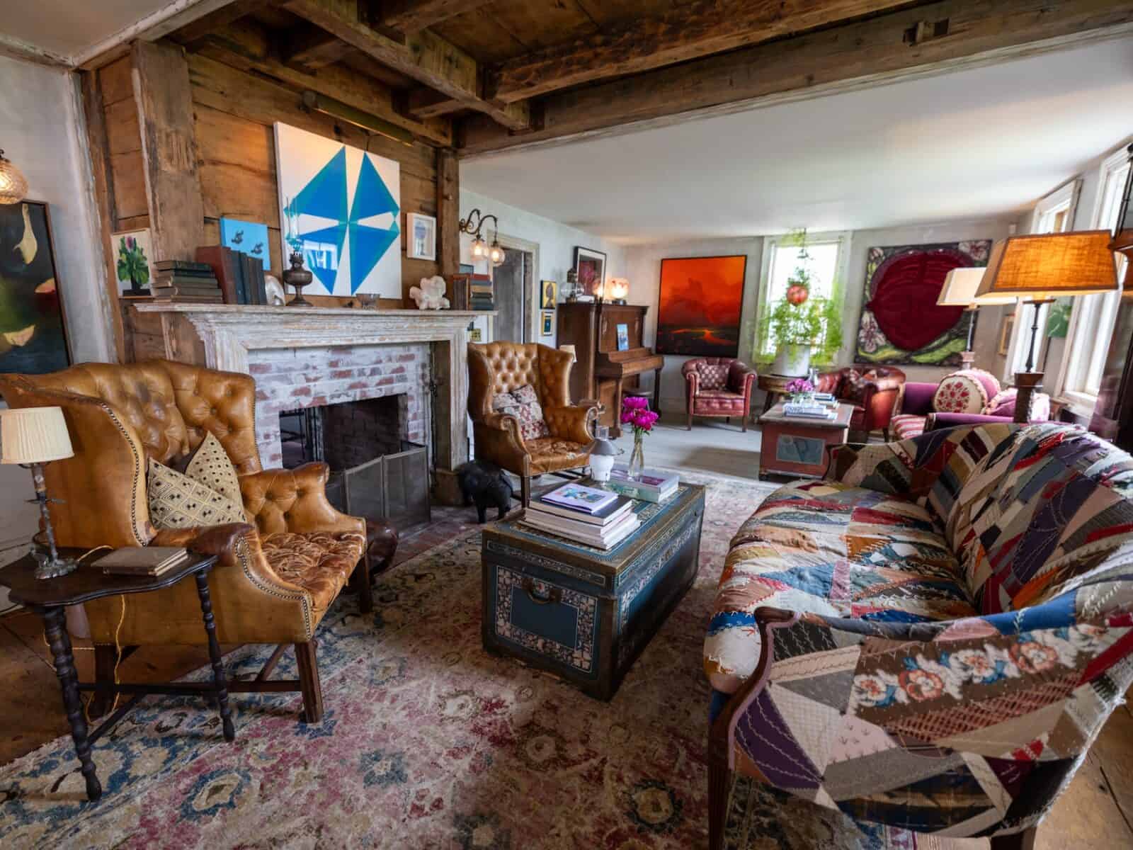 A wide view of a spacious, eclectic living room inside a historic Provincetown house featuring exposed wooden ceiling beams, weathered leather tufted wingback chairs, a patchwork fabric sofa, an antique trunk coffee table, and an open brick fireplace adorned with original artwork.
