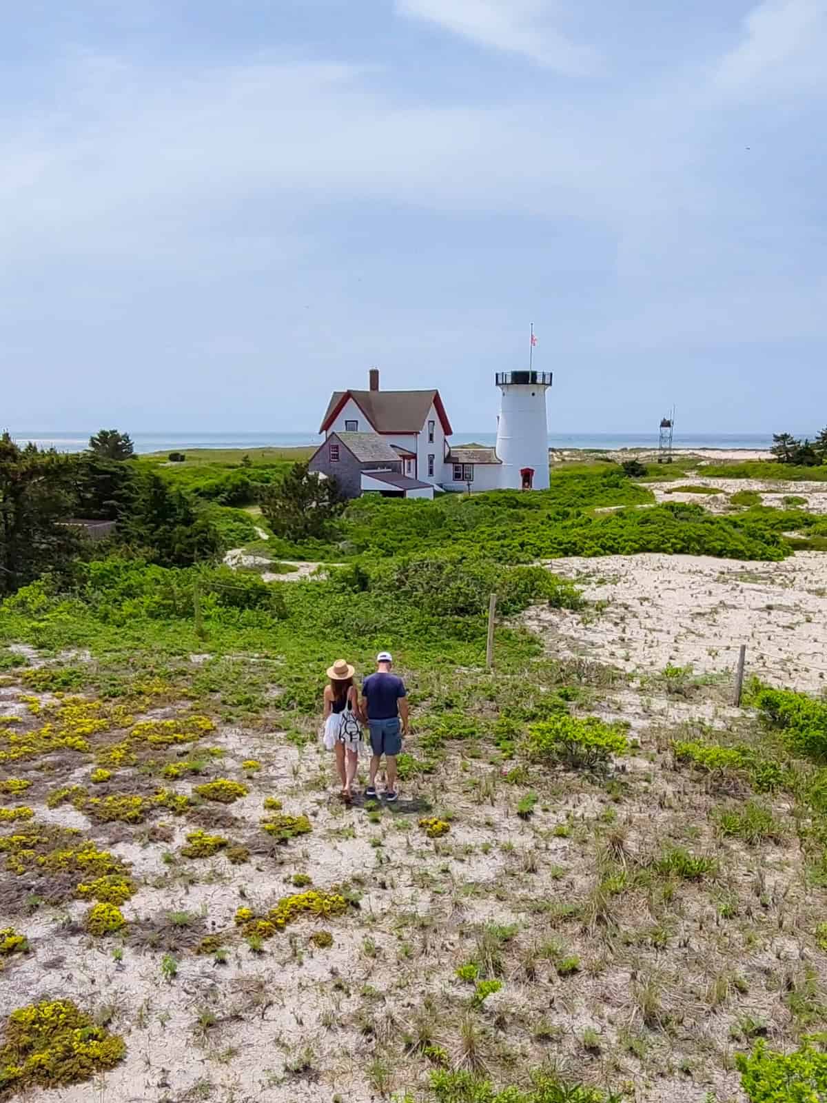 An aerial-style shot looking down at Laura and Luca walking hand-in-hand along a sandy beach trail through dune vegetation toward the historic Stage Harbor Lighthouse and keeper's dwelling in Chatham, Cape Cod.