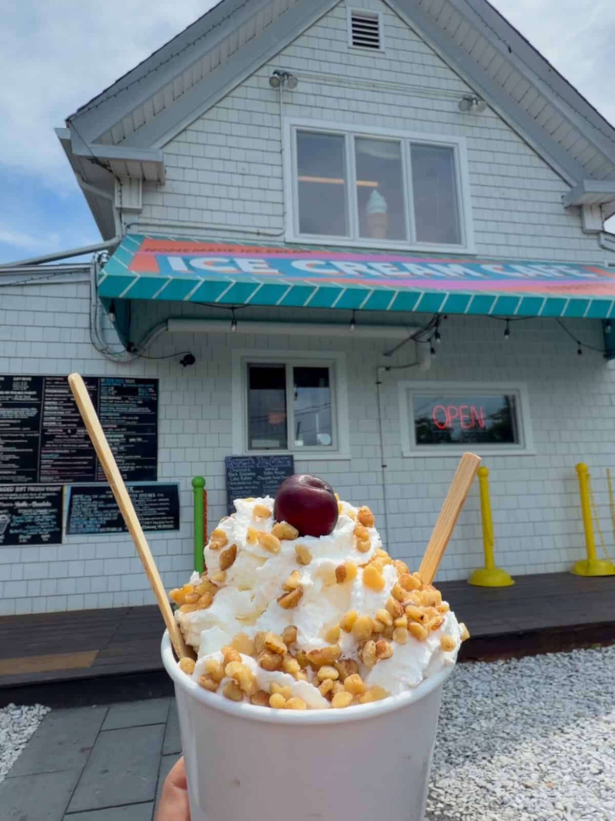 Laura up a massive ice cream sundae layered with thick whipped cream, chopped walnuts, and a single dark cherry on top, framed in front of the white-shingled storefront and colorful awning of the Ice Cream Cafe in Orleans