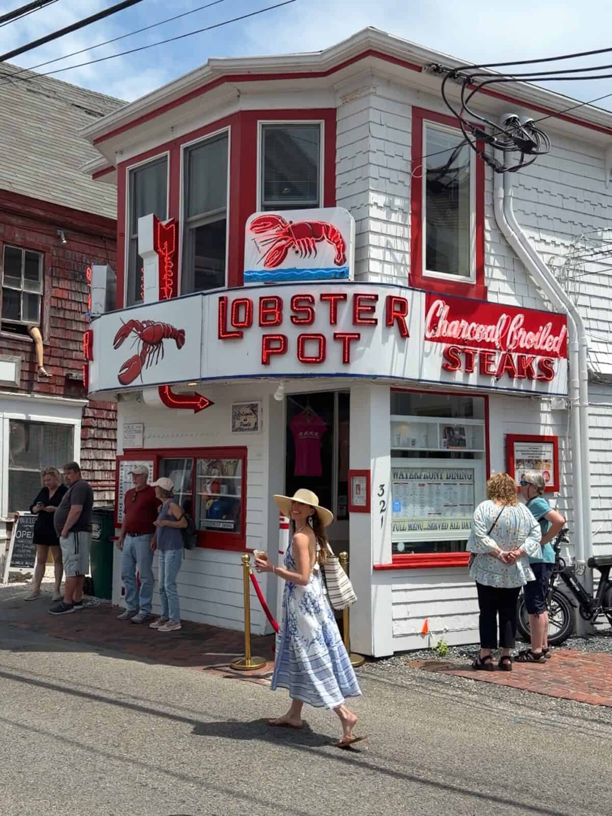 Laura wearing a wide-brimmed straw hat and a blue-and-white maxi dress walks past the iconic red-and-white neon storefront of The Lobster Pot restaurant on Commercial Street in Provincetown.