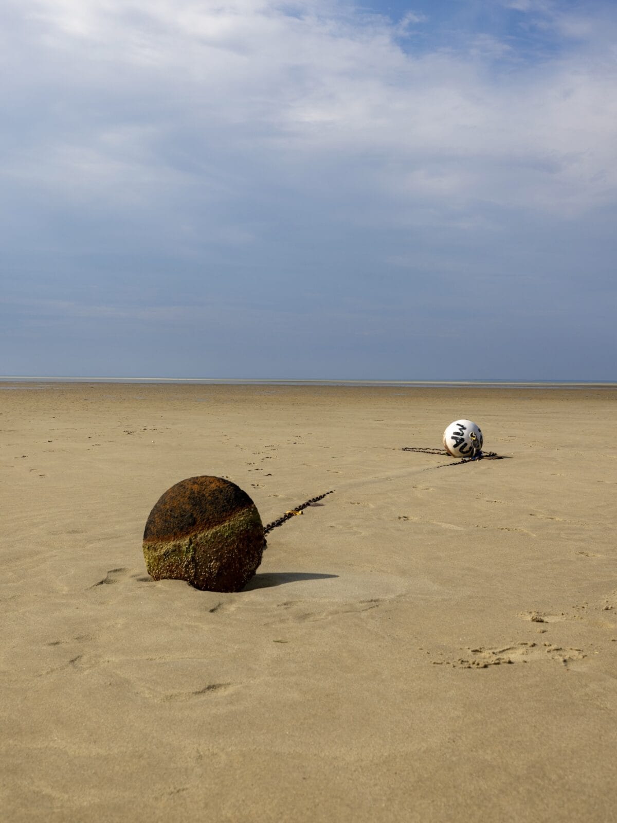 An open landscape shot of the Brewster Flats at low tide, featuring a rusted iron anchor ball in the foreground connected by a long heavy chain to a white plastic mooring buoy resting on dry sand under a blue sky.