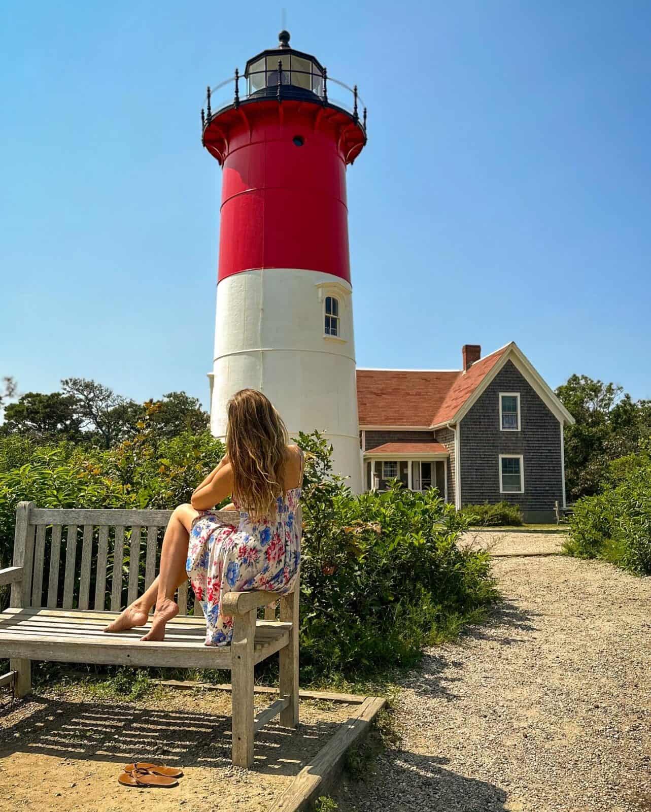 Laura wearing a white floral maxi dress sits on a wooden park bench, looking up at the iconic red-and-white Nauset Lighthouse in Eastham, with a historic cedar-shingled keeper's home and a gravel pathway visible in the background under a clear blue sky.