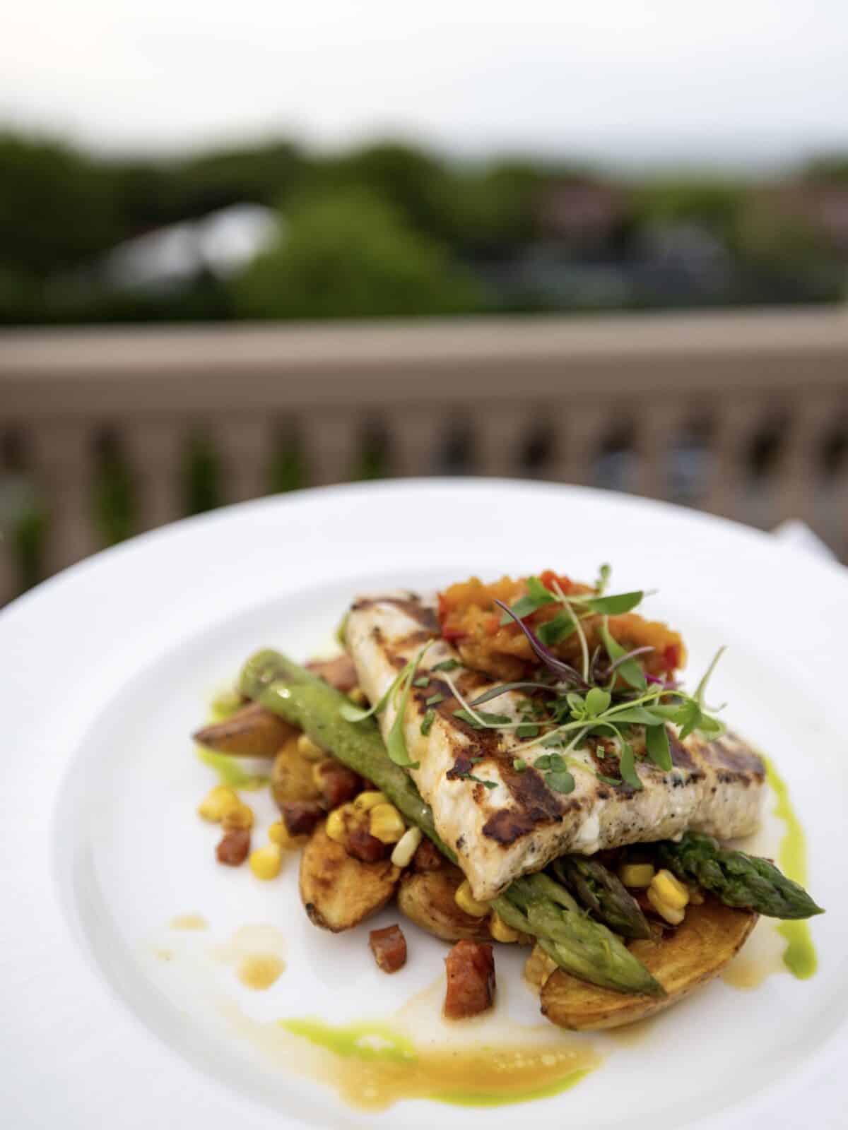 A close-up view of a perfectly grilled swordfish steak topped with fresh microgreens and tomato relish, layered over grilled asparagus, roasted fingerling potatoes, and sweet corn with bacon bits at Ocean Terrace.