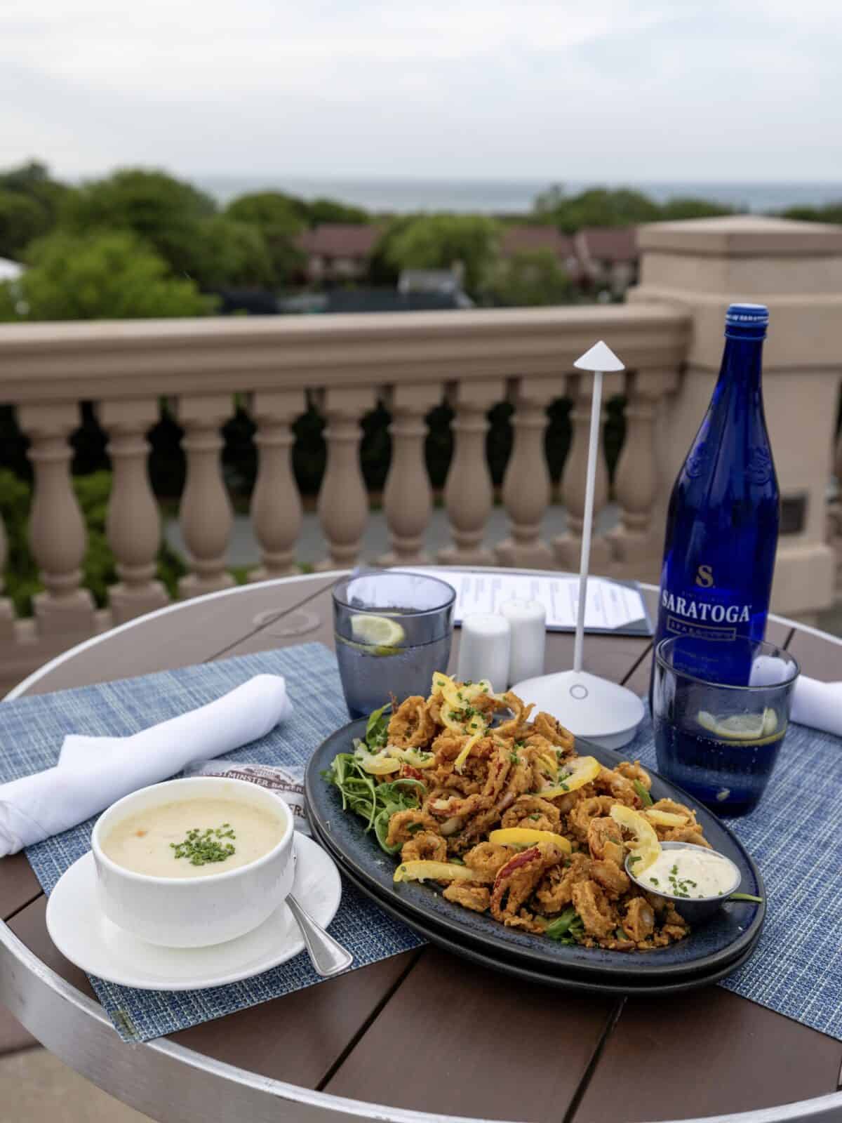 A white tabletop setup on an outdoor balcony at Ocean Terrace showcasing a bowl of creamy New England clam chowder topped with chives, a large platter of crispy fried calamari with lemon wheels, and a blue bottle of Saratoga sparkling water.