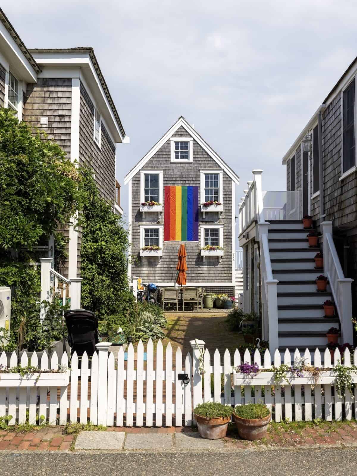 A charming New England classic shingle-style home in Provincetown framed by a white picket fence, featuring a large vibrant rainbow pride flag hanging prominently on its front facade between multi-pane windows.