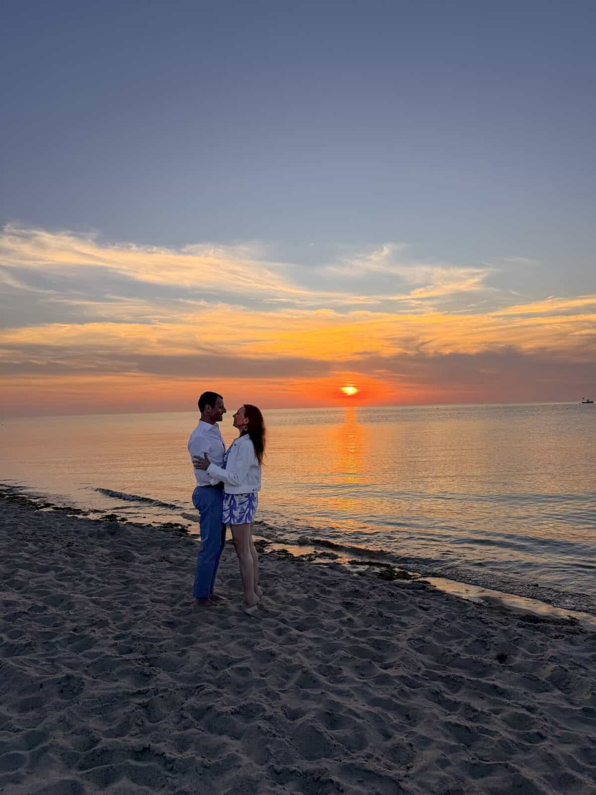 A romantic portrait of Laura and Luca embracing on the sandy shoreline of the private beach at Ocean Edge Resort as the sun dips below the horizon, casting a fiery orange and yellow glow across the calm water.