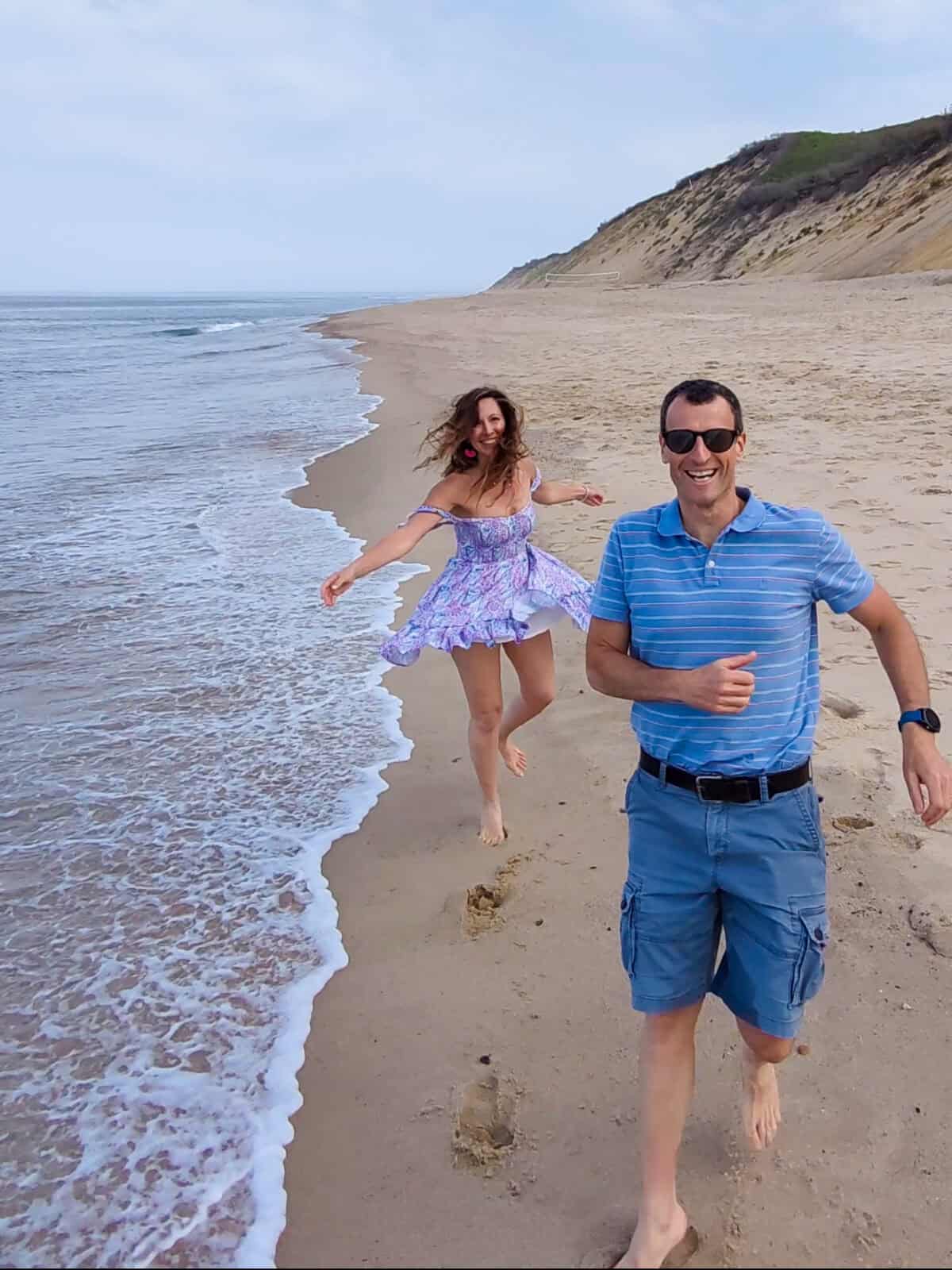 A joyful couple runs barefoot along the wet sand at the ocean's edge at Cahoon Hollow Beach in Wellfleet, with Laura laughing in a flowing purple sundress and Luca in a striped polo shirt, backdropped by a towering sand dune