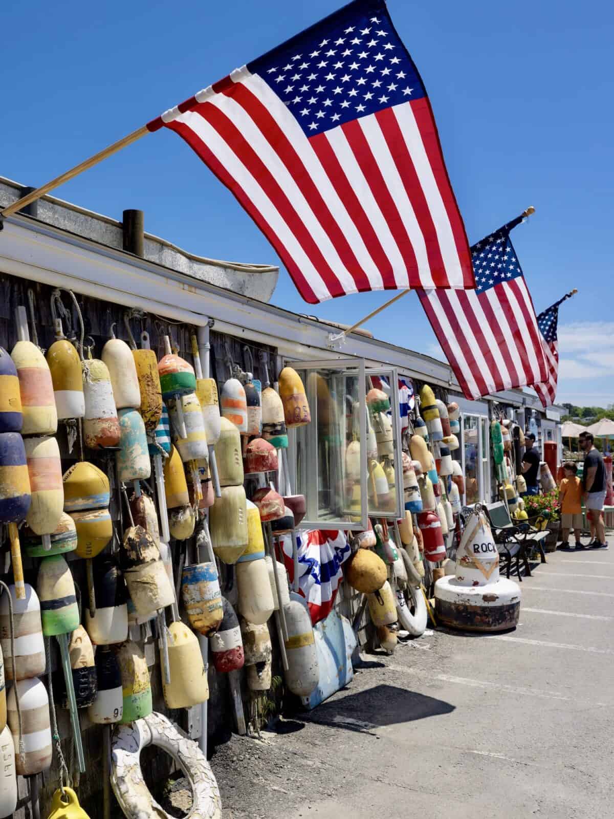 A vibrant outdoor wall of a weathered shingle building completely covered in hundreds of colorful, painted antique lobster buoys beneath three large American flags flying against a bright blue sky.