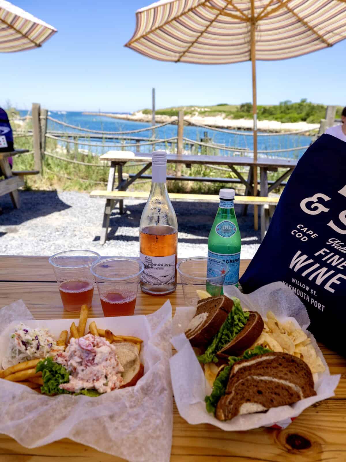 A wooden picnic table under a striped umbrella features a fresh lobster roll with fries and coleslaw, a turkey sandwich, a bottle of chilled rosé from Dunn & Sons Wine, and a green S.Pellegrino bottle overlooking the blue waters of Sesuit Harbor.